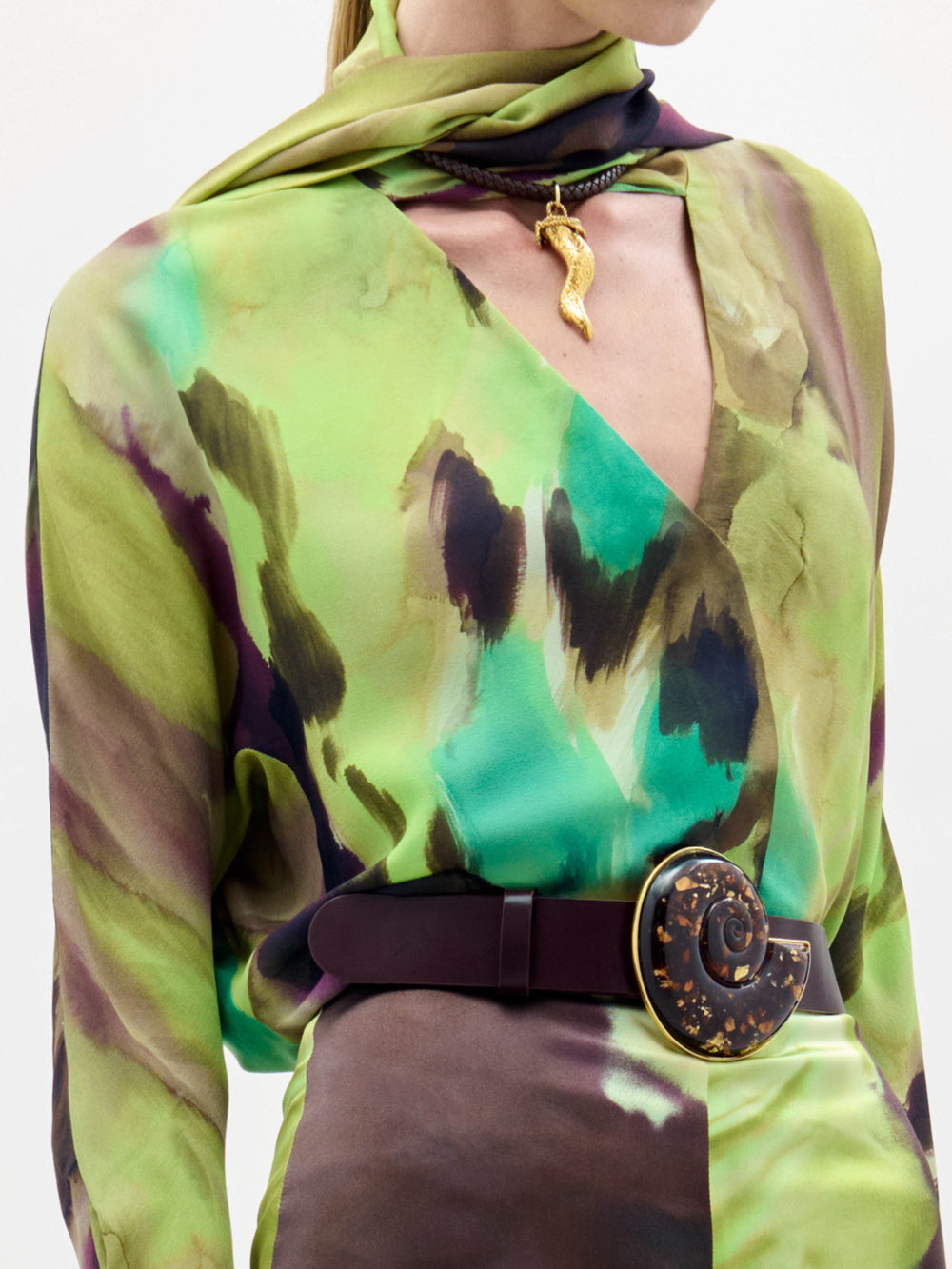 A woman stands against a plain white background wearing the Jaboca Blouse—a green, yellow, and brown tie-dye top styled with a wide belt and a scarf draped around her neck.