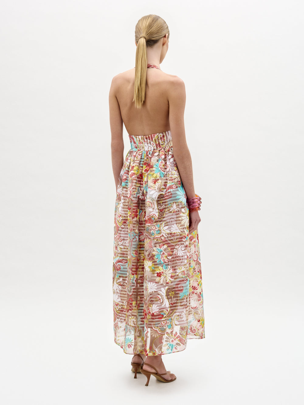 A woman faces away from the camera in the SVT DESIGNS Kairi Dress, a strapless, ankle-length floral pastel A-line midi. She wears brown heeled sandals, a pink bracelet, and has a low ponytail against a plain white background.