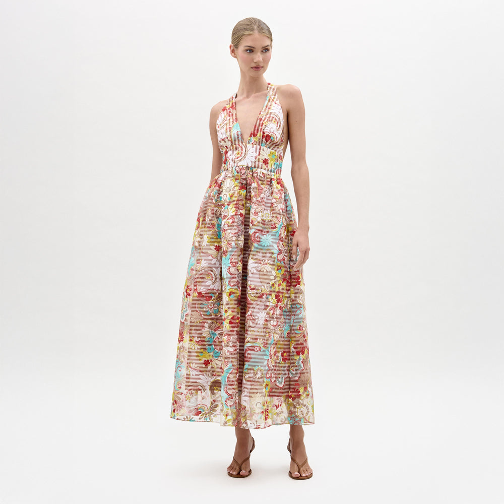 A woman poses against a white background wearing the Kairi Dress by SVT DESIGNS—a sleeveless, floor-length piece made from Italian fabric with a deep V-neckline and colorful intricate patterns, styled with brown sandals.
