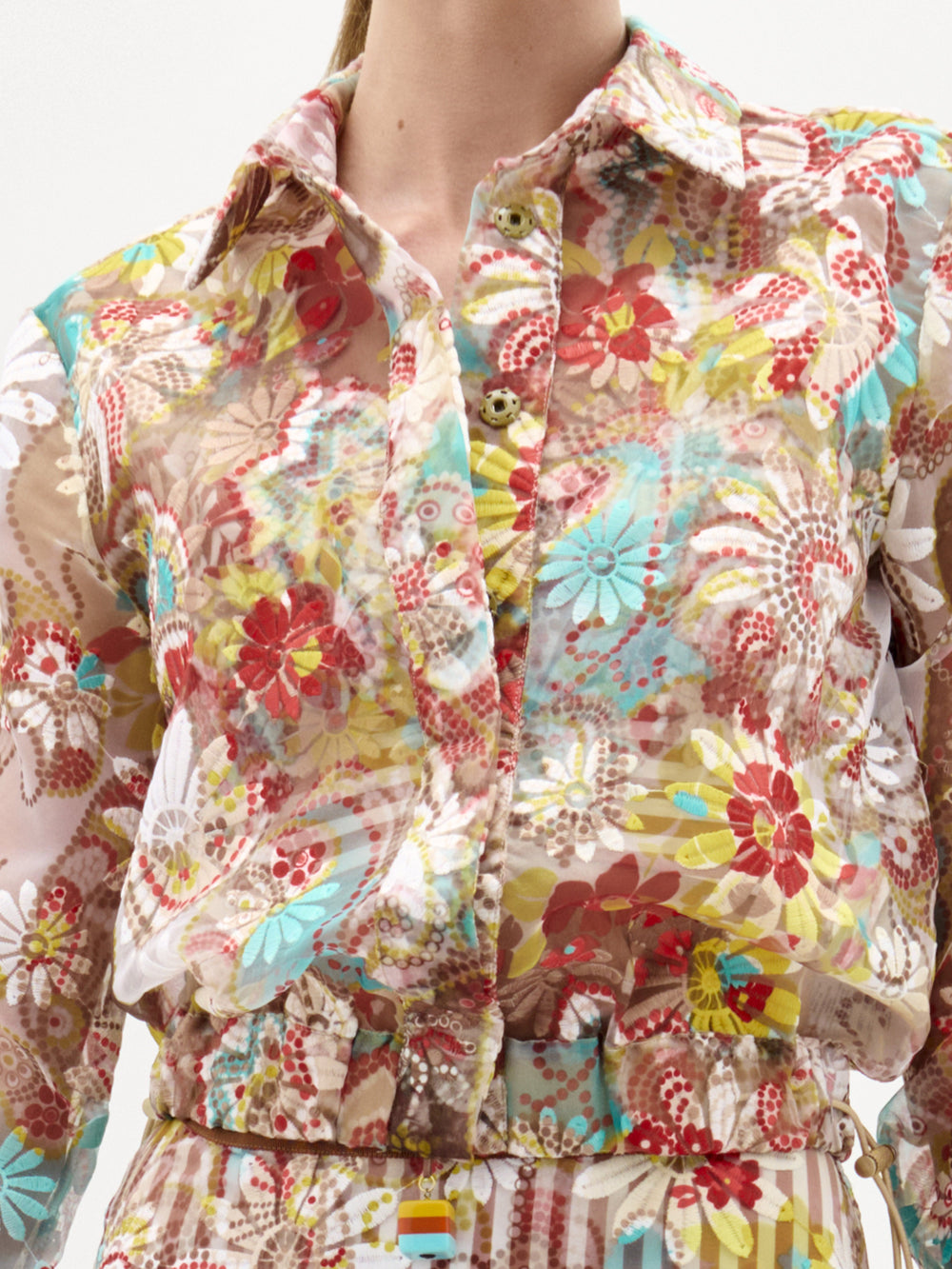 A person wearing the Katniss Jacket by SVT DESIGNS, a sheer, long-sleeved piece with a colorful floral pattern in red, yellow, blue, and beige. Made from Italian fabric, it features front buttons and a collar.