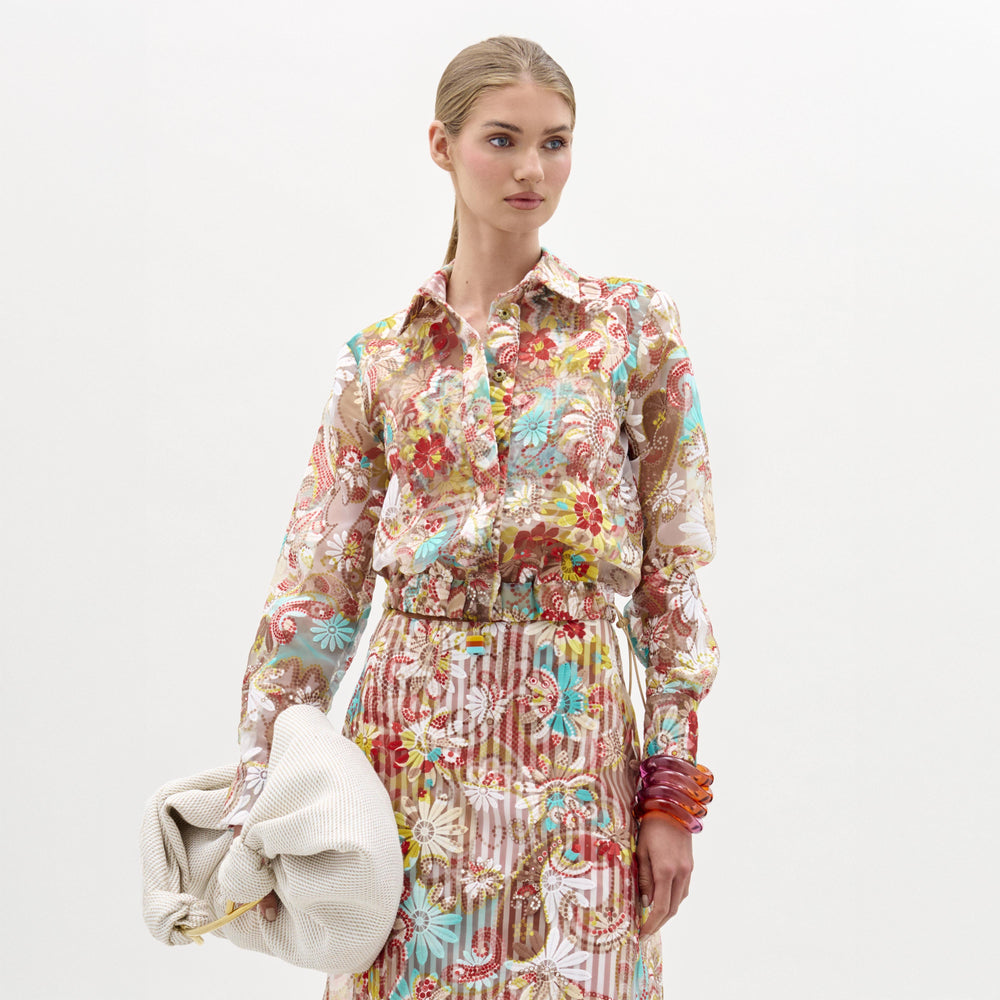 A woman with blonde hair pulled back wears SVT DESIGNS’ Katniss Jacket over a sheer floral blouse and matching skirt, accessorized with a large white clutch and colorful bangles, standing against a plain white background.