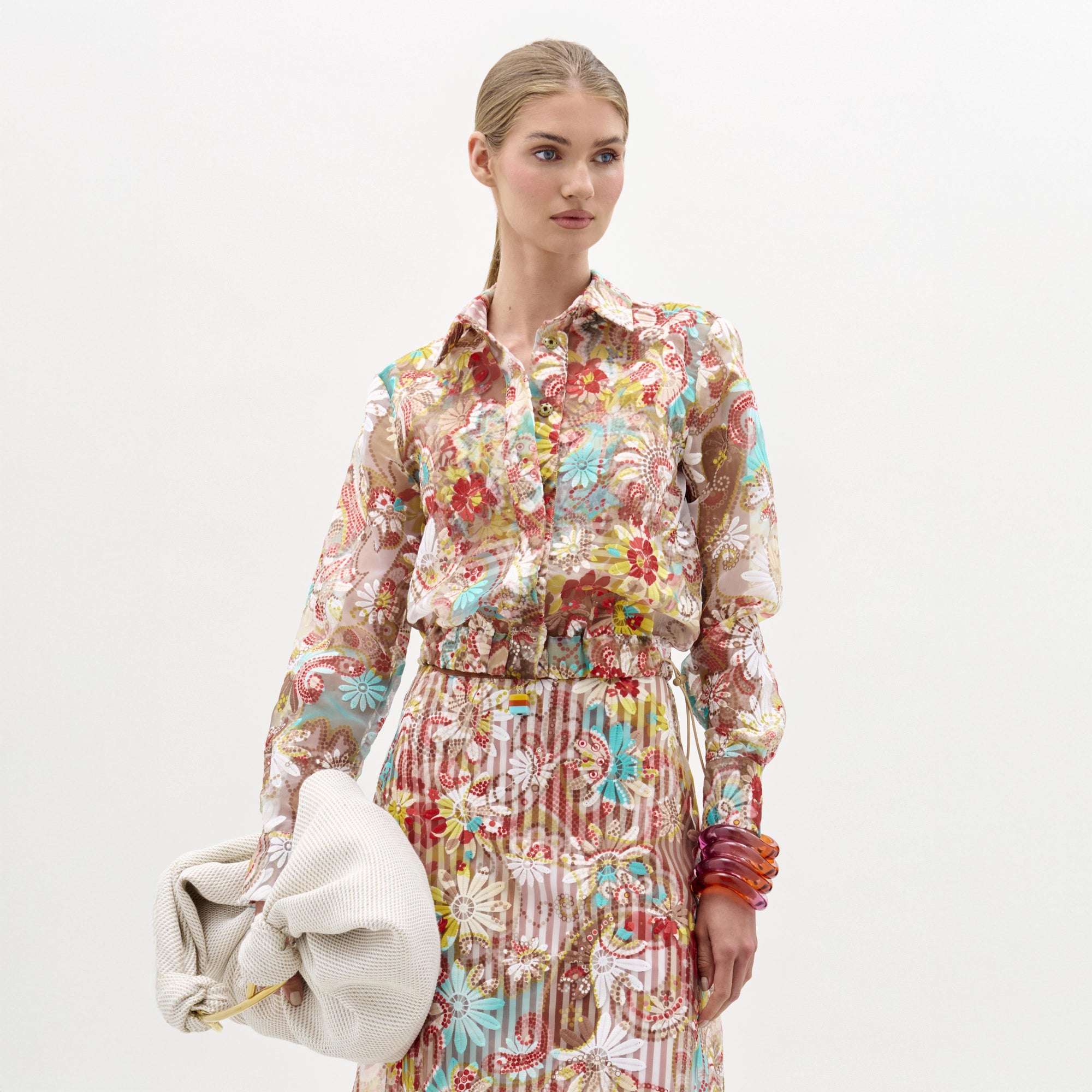 A woman with blonde hair pulled back wears SVT DESIGNS’ Katniss Jacket over a sheer floral blouse and matching skirt, accessorized with a large white clutch and colorful bangles, standing against a plain white background.