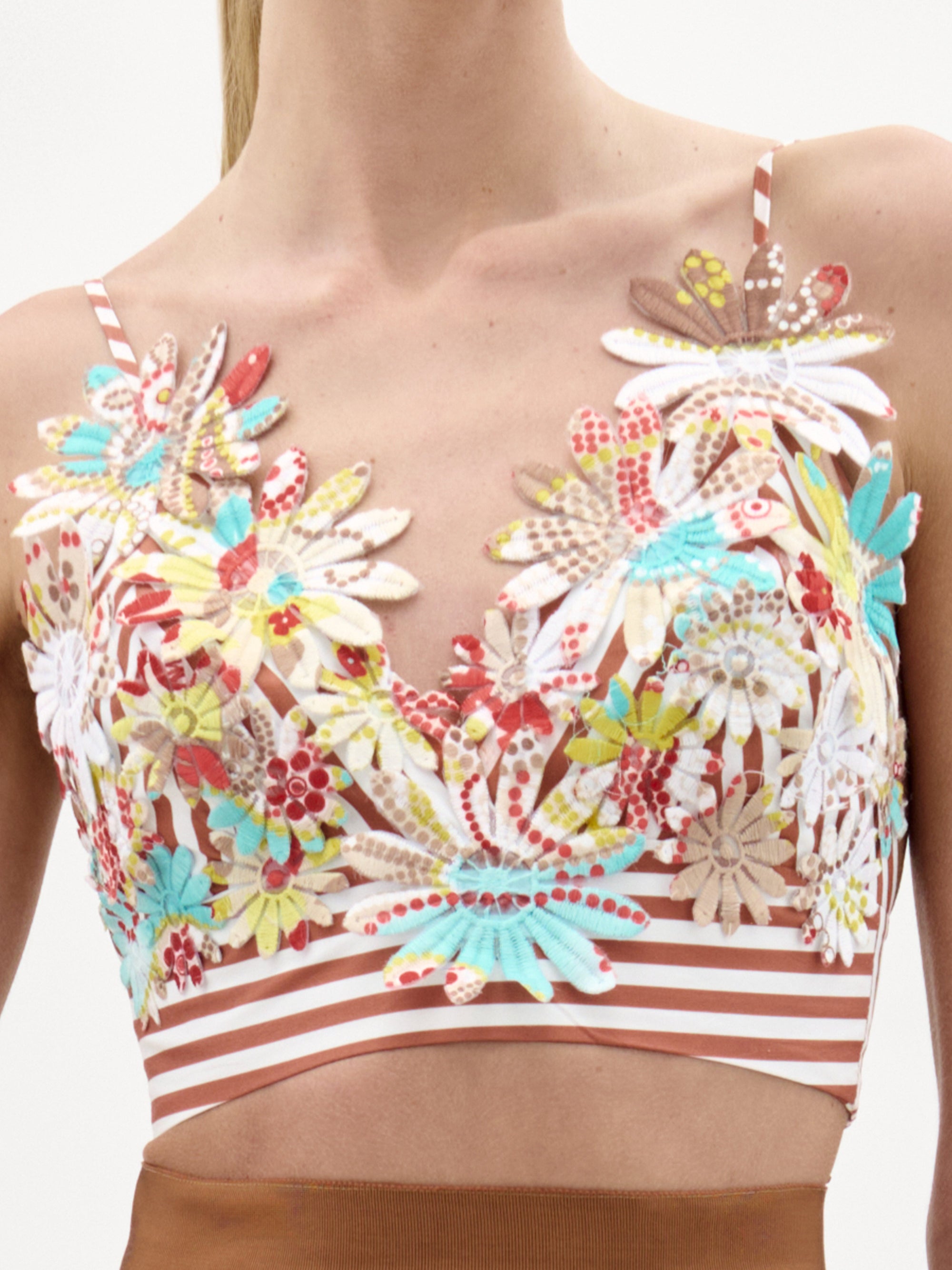 A person wears the Ibbie Top by SVT DESIGNS, a floral embroidered cropped top with colorful flowers on sheer fabric and a brown and white striped band at the hem; their lower face and torso are visible.