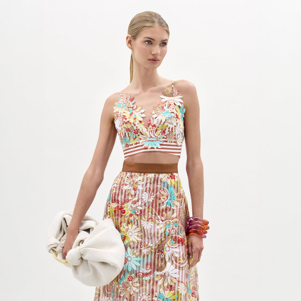 A woman stands against a plain background wearing the Ibbie Top by SVT DESIGNS with a matching maxi skirt, holding a large textured clutch and chunky red bracelets, her hair pulled back neatly.