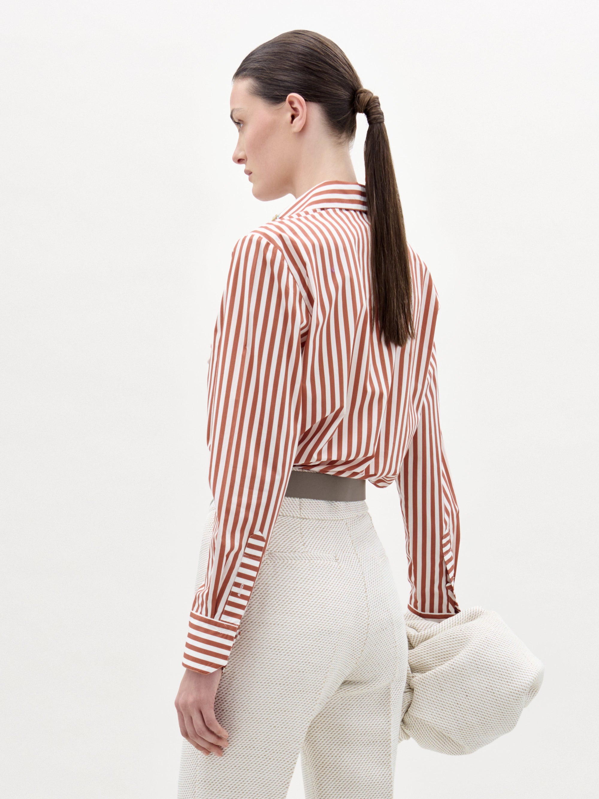 A woman with straight brown hair in a low ponytail wears the SVT DESIGNS Karole Blouse, a red and white striped button-down, tucked into high-waisted textured white pants and holds a light-colored textured item in her left hand.