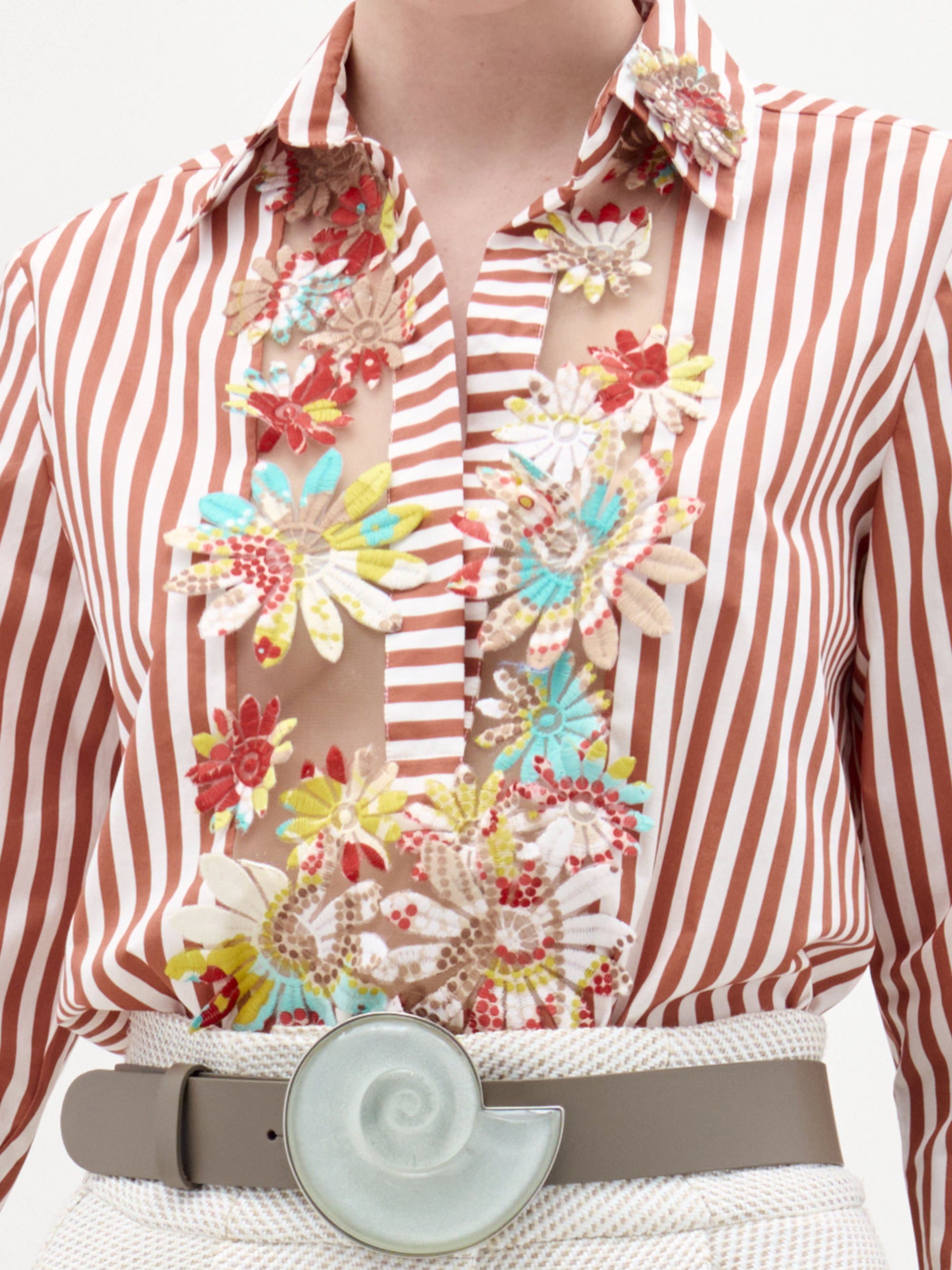 A person wears the Karole Blouse by SVT DESIGNS, a brown and white striped button-down with colorful floral embroidery on fine Italian fabric, tucked into light pants and styled with a wide belt featuring a nautilus shell buckle.
