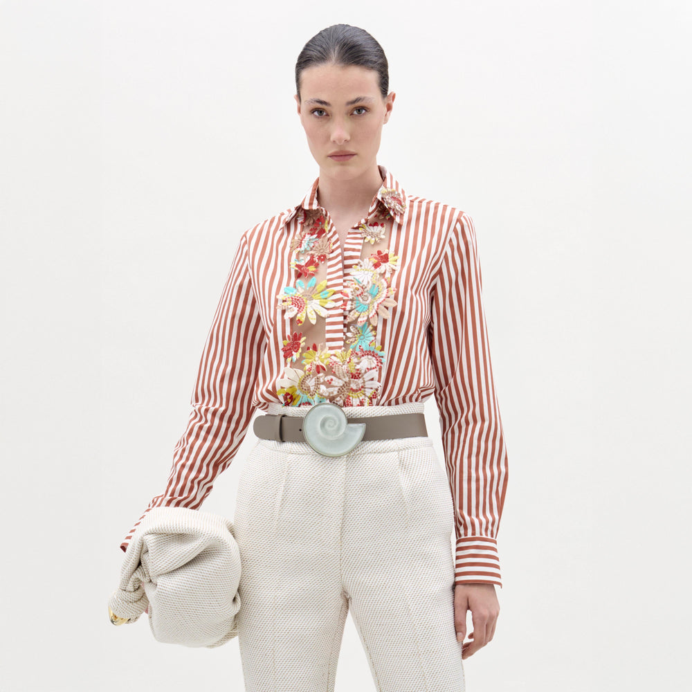 A woman stands against a plain background wearing the Karole Blouse by SVT DESIGNS, a floral embroidered, striped button-down crafted from Italian fabric. She pairs it with high-waisted textured white pants, a wide round-buckle belt, and holds a light jacket.