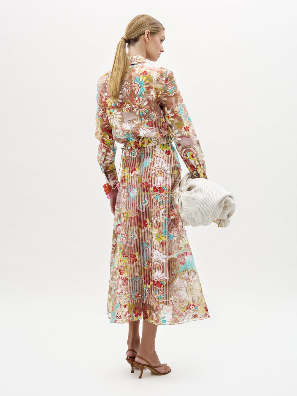 A woman faces away, wearing the Nelida Skirt by SVT DESIGNS, a long sheer floral piece in Italian fabric with red, green, and blue patterns. She holds an off-white textured clutch, wears brown heeled sandals, and her hair is in a low ponytail.