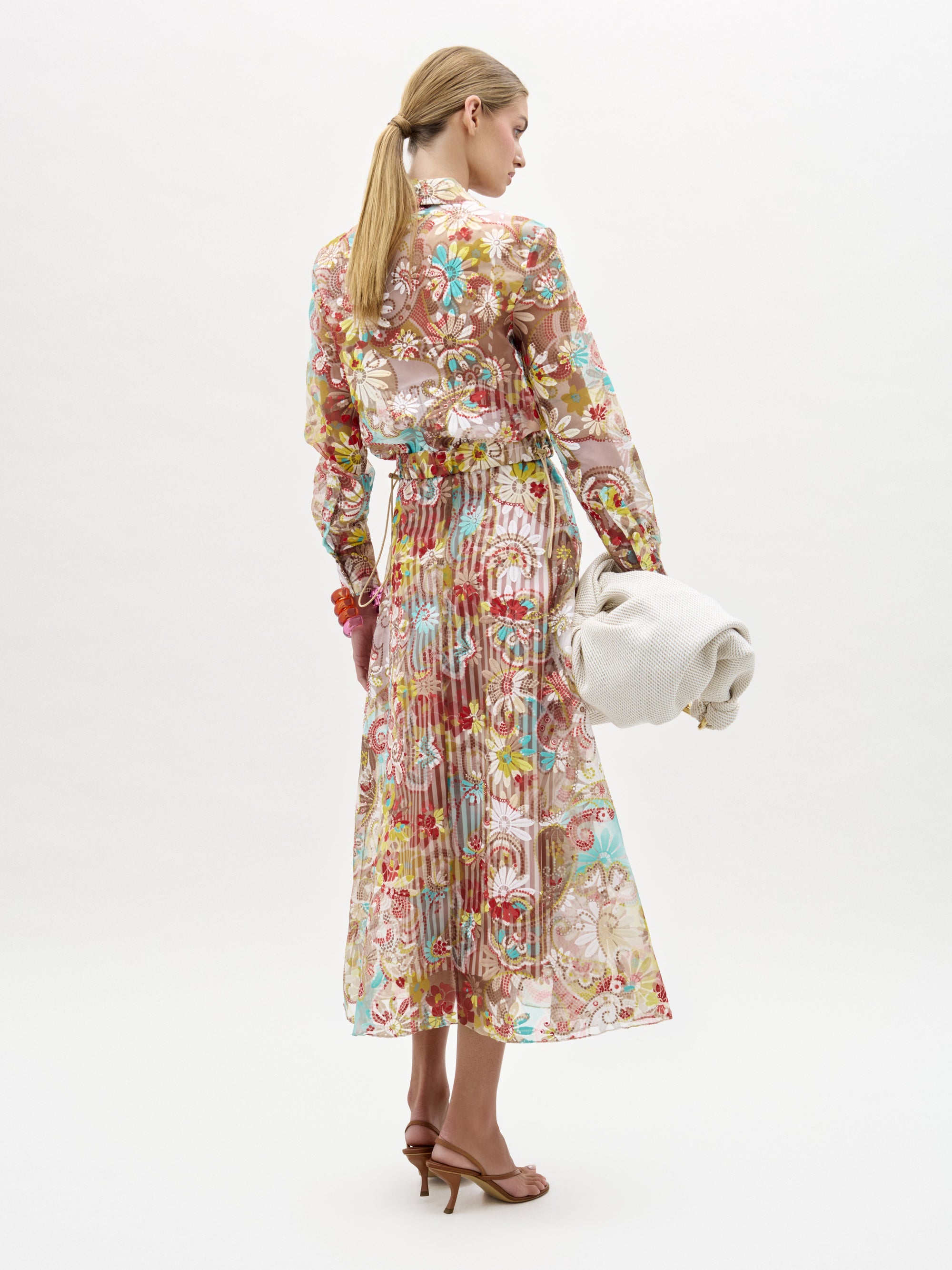 A woman faces away, wearing the Nelida Skirt by SVT DESIGNS, a long sheer floral piece in Italian fabric with red, green, and blue patterns. She holds an off-white textured clutch, wears brown heeled sandals, and her hair is in a low ponytail.