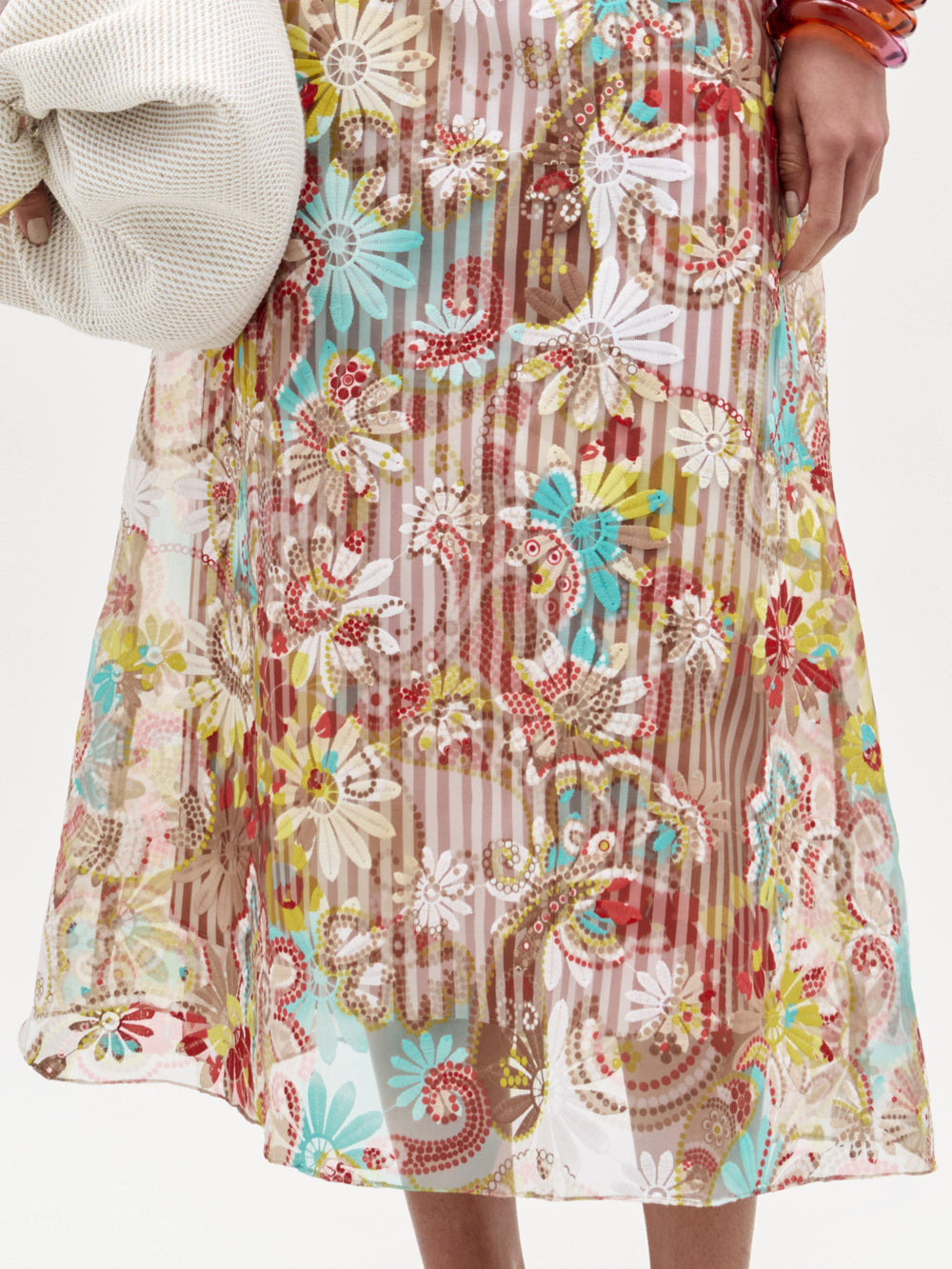 A person wearing the SVT DESIGNS Nelida Skirt—a high-waisted, sheer midi skirt featuring a colorful floral and paisley pattern over a neutral slip—holds a light-colored textured garment in their left arm.