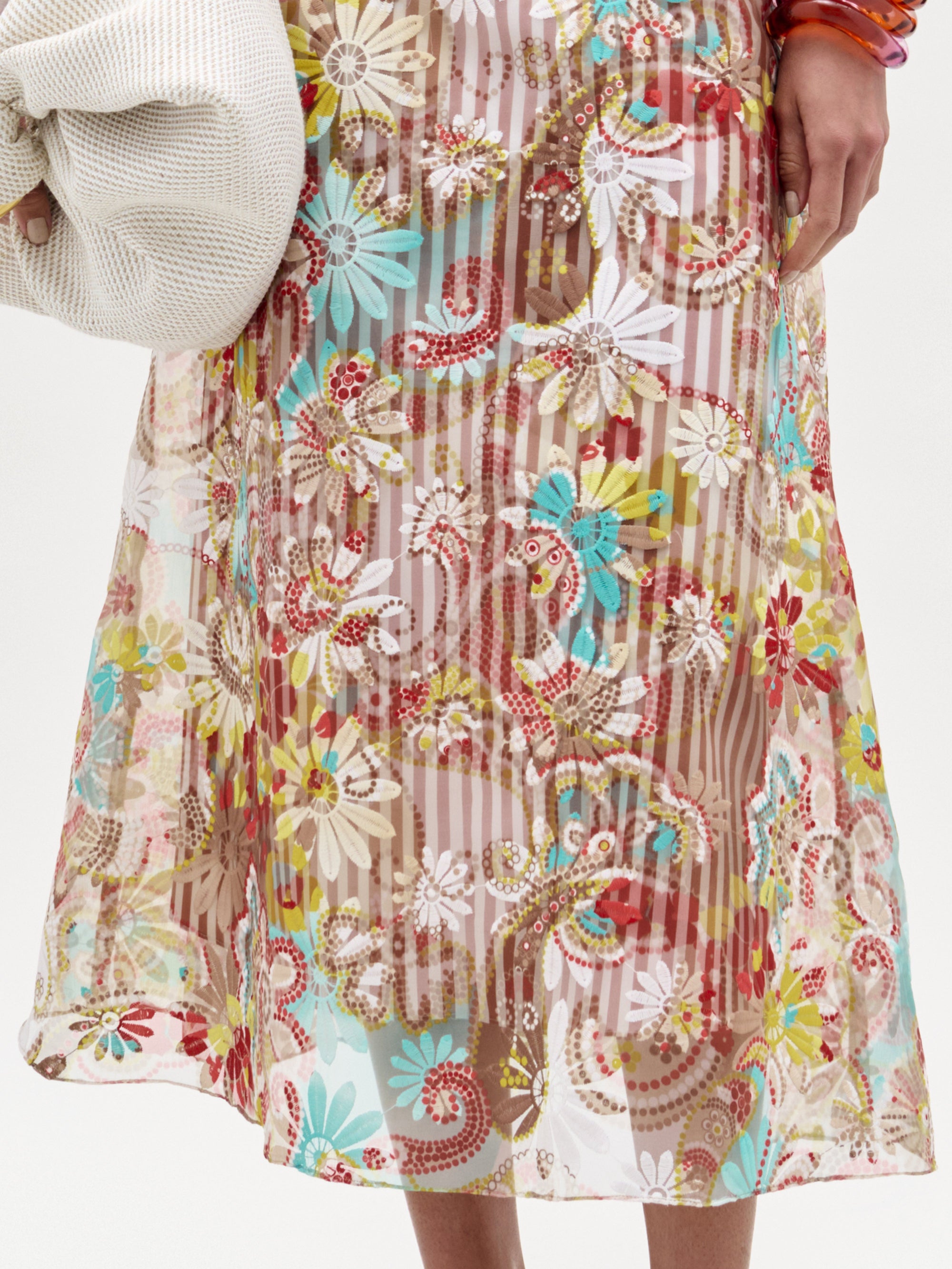 A person wearing the SVT DESIGNS Nelida Skirt—a high-waisted, sheer midi skirt featuring a colorful floral and paisley pattern over a neutral slip—holds a light-colored textured garment in their left arm.