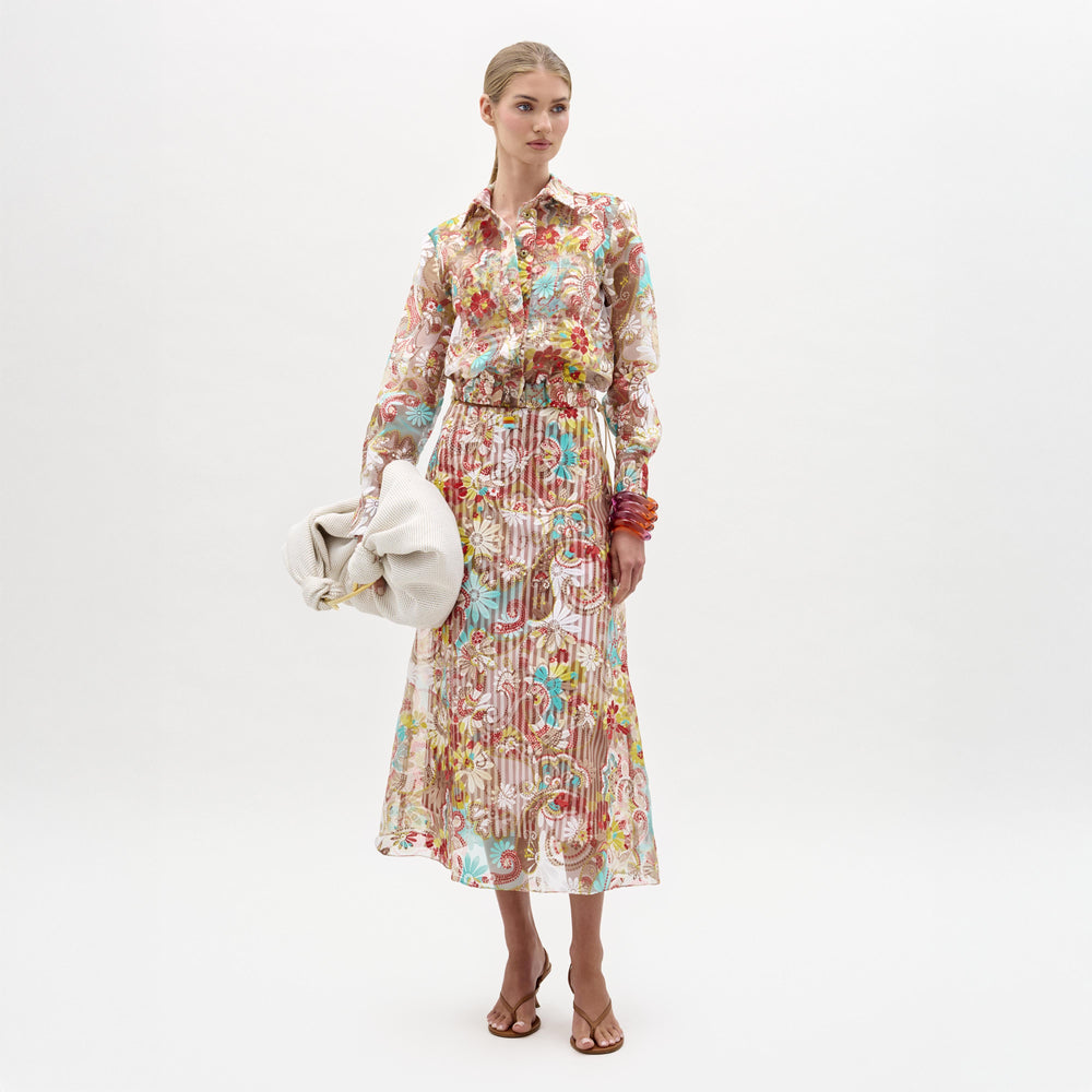 A woman stands against a white background wearing the Nelida Skirt by SVT DESIGNS, a high-waisted midi made from Italian fabric, paired with a matching sheer floral blouse, large beige bag, brown heeled sandals, and colorful bangles.