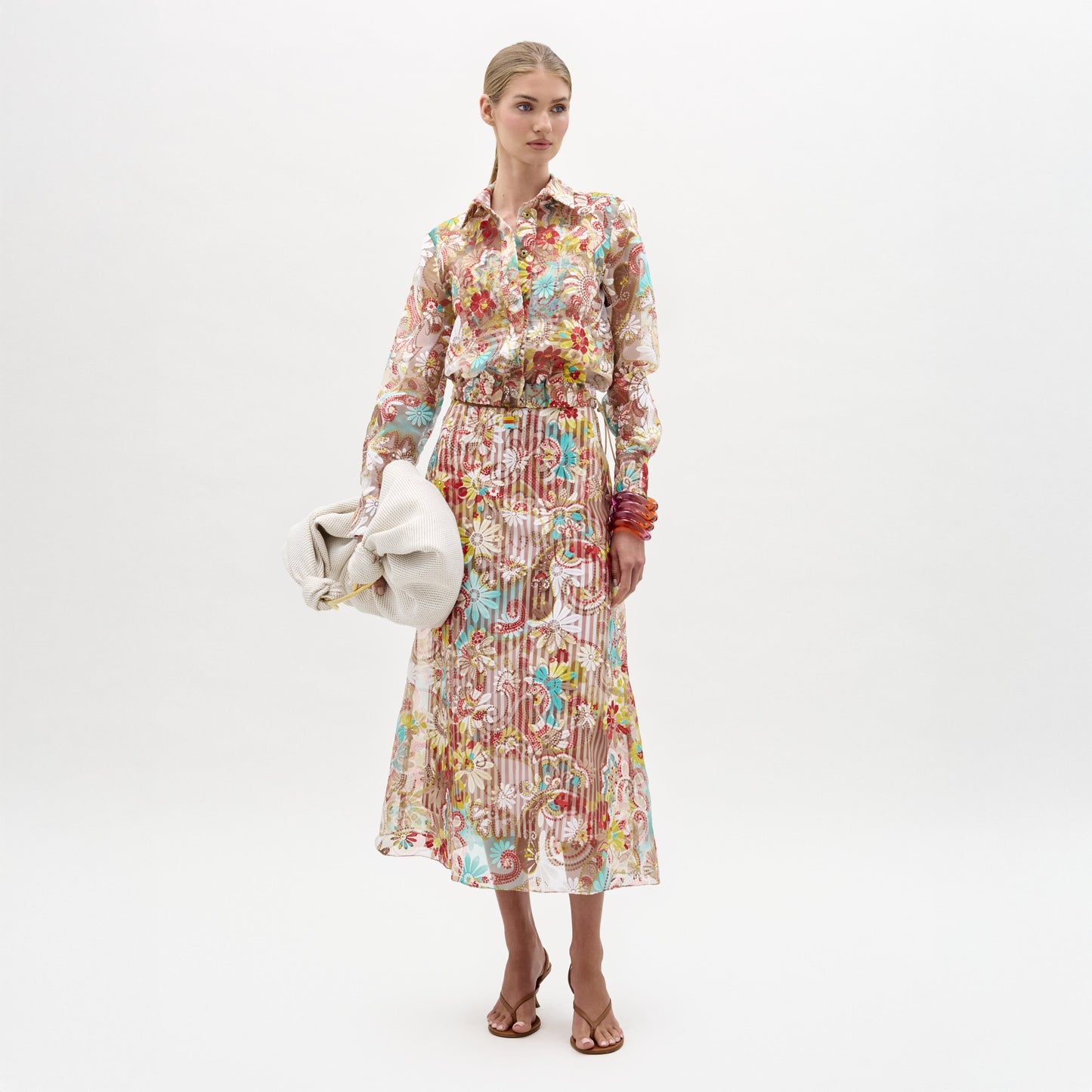 A woman stands against a white background wearing the Nelida Skirt by SVT DESIGNS, a high-waisted midi made from Italian fabric, paired with a matching sheer floral blouse, large beige bag, brown heeled sandals, and colorful bangles.