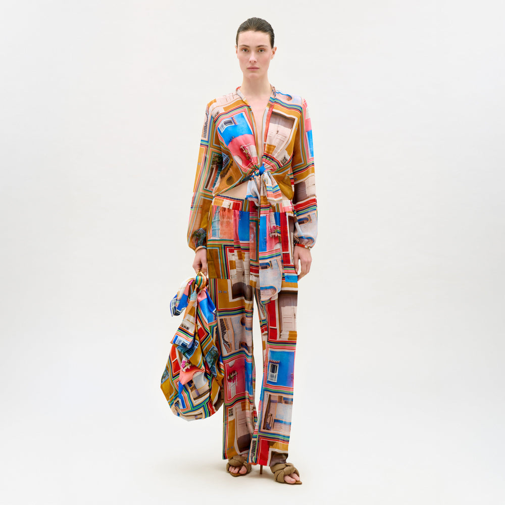 A woman stands against a white background wearing the Nerea Pant, a colorful Italian fabric outfit with window patterns, holding a matching bag. She has brown sandals and her hair is pulled back.