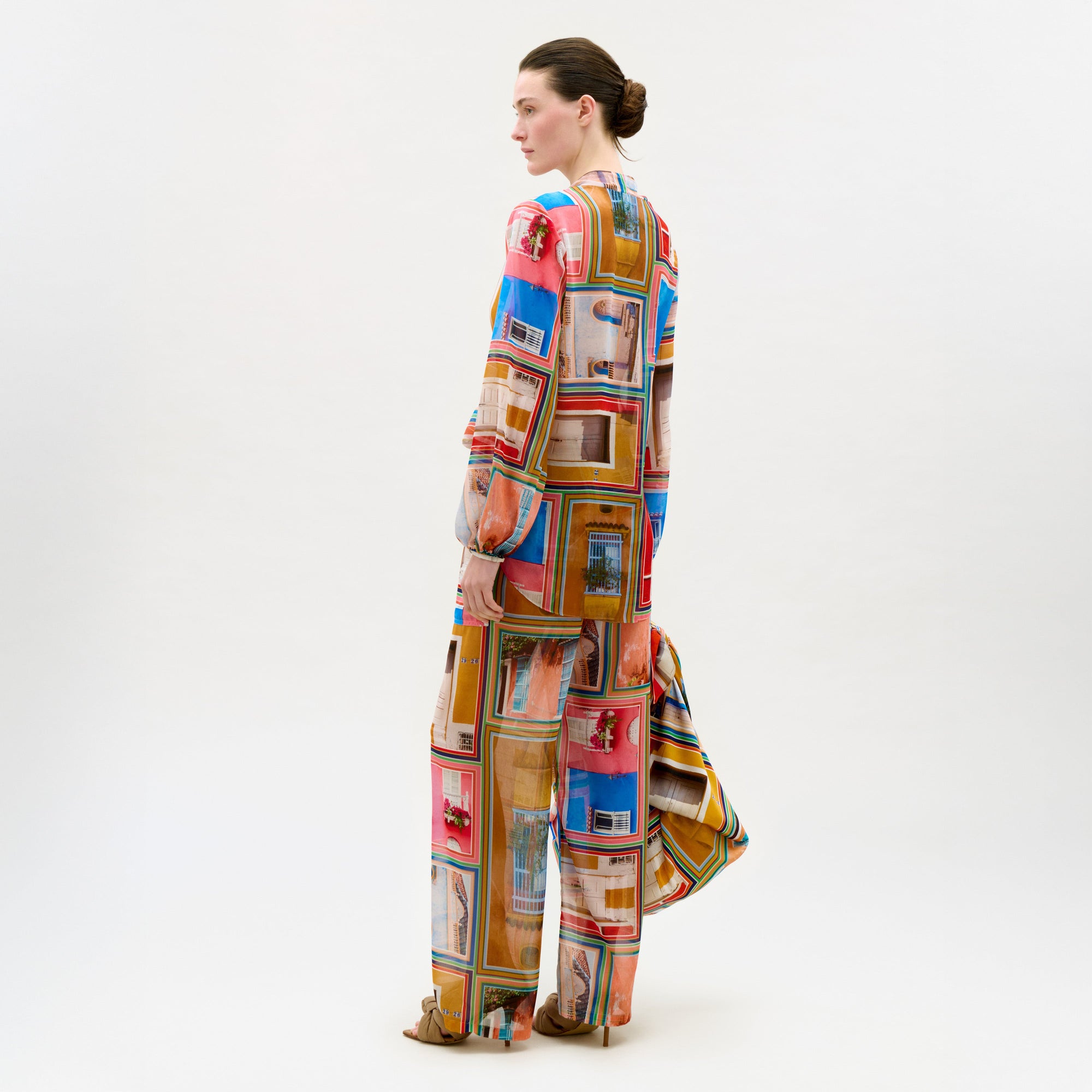 A woman with dark hair in a bun models the Nerea Pant—loose-fitting, long-sleeve chiffon pants with an elastic waistband and colorful geometric photo-print—standing sideways against a plain white background.