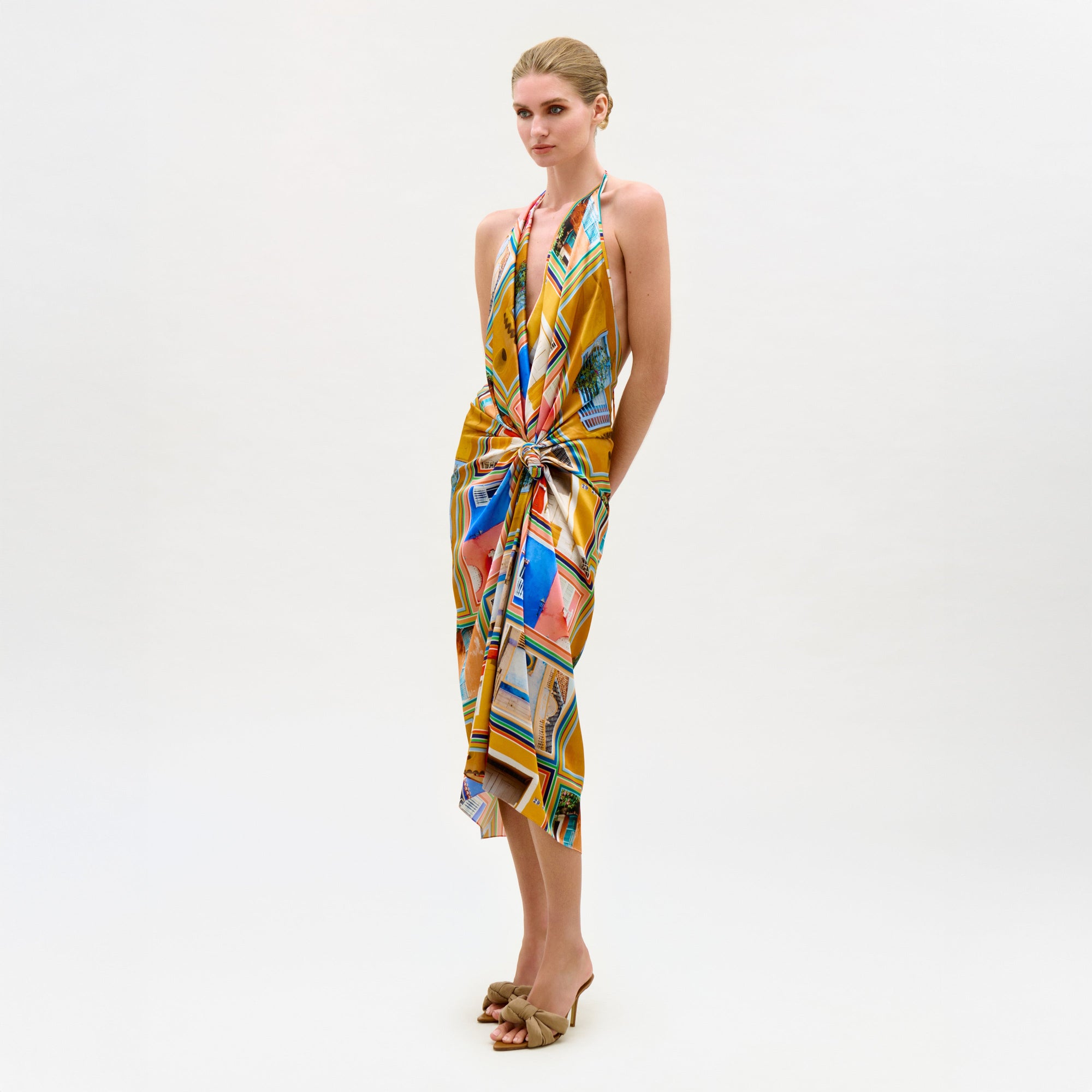 A woman stands against a plain white background, wearing the Guadalupe Dress—a colorful silk midi with a halter neckline and knotted waist—paired with tan heeled sandals. Her hair is pulled back, and her expression is neutral.