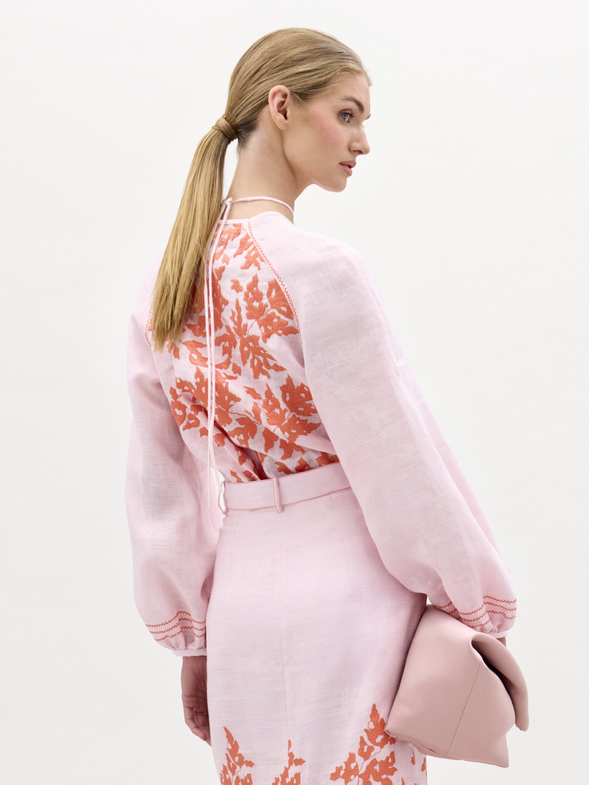 A blonde woman in a low ponytail is seen from the side, wearing the SVT DESIGNS Molveno Blouse with a V neckline and orange floral embroidery, styled with a matching skirt and blush pink clutch against a plain white background.