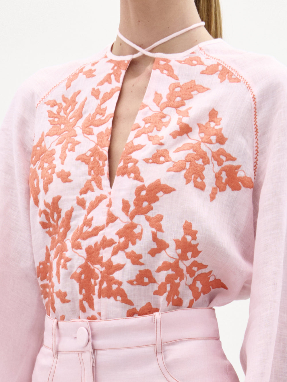 A person wears the SVT DESIGNS Molveno Blouse—light pink Italian linen with orange leaf embroidery, a front keyhole neckline, and thin neck strap—paired with high-waisted light pink pants.