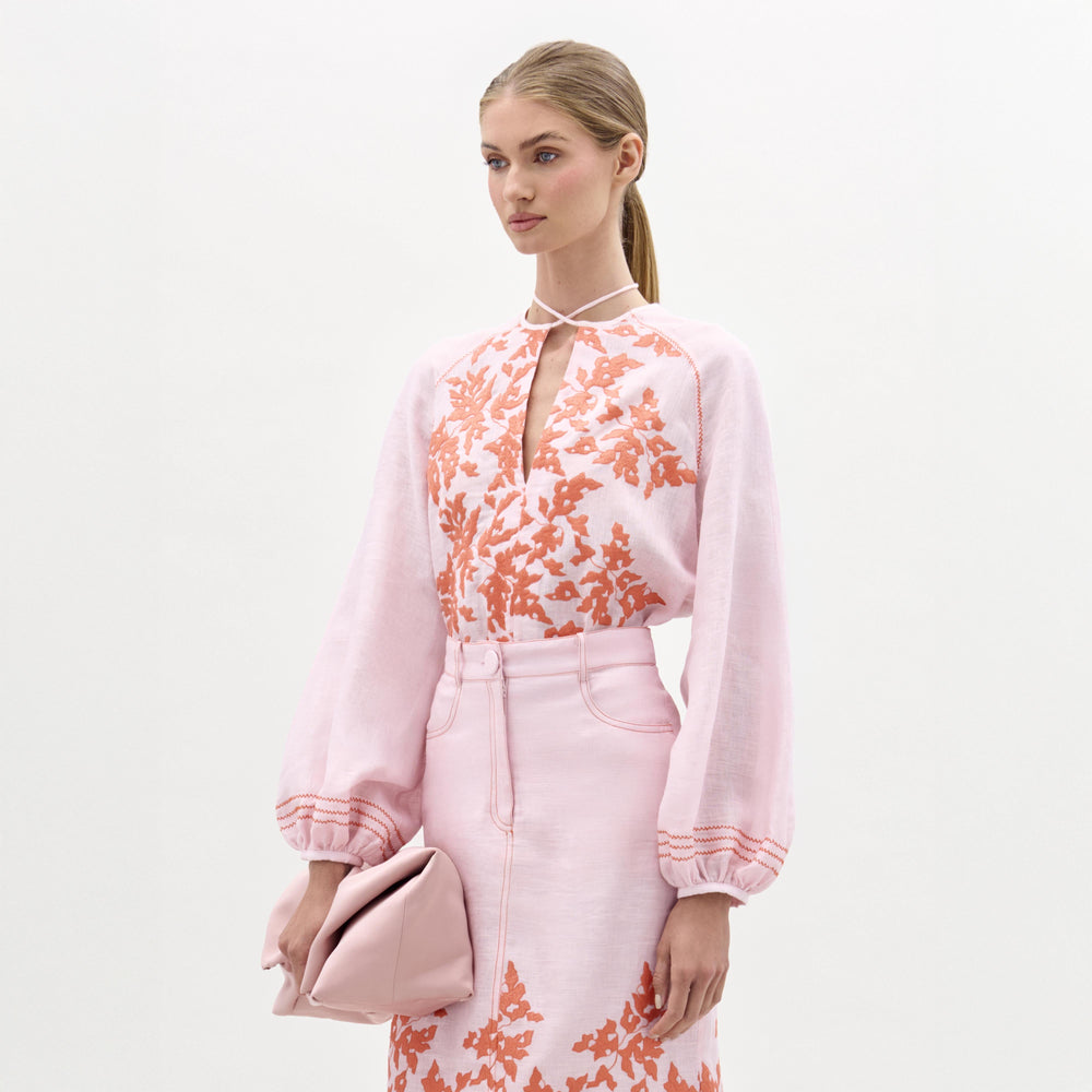 A woman stands against a plain background wearing the Molveno Blouse by SVT DESIGNS—an embroidered light pink linen top with orange floral patterns—and holds a soft pink clutch. Her hair is styled in a low ponytail.