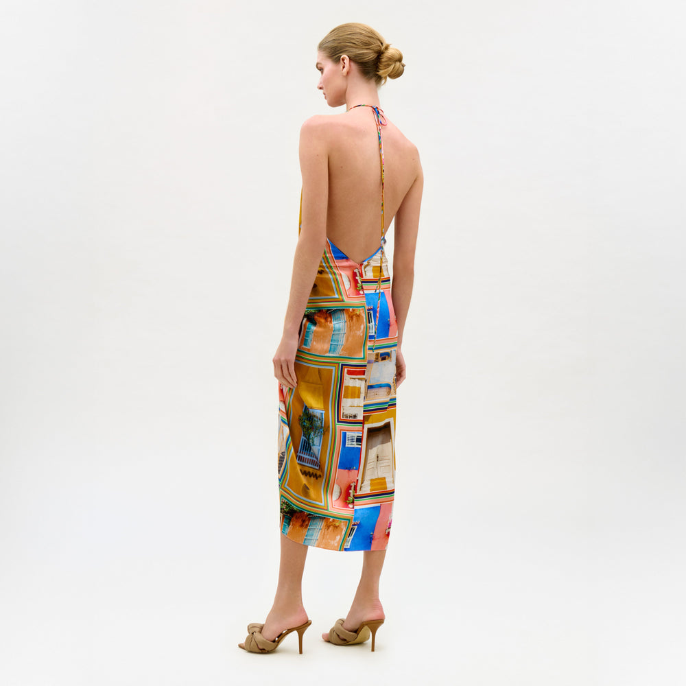 A woman with blonde hair in a neat bun wears the colorful silk Guadalupe Dress with a halter neckline and beige high-heeled sandals, standing against a plain white background.