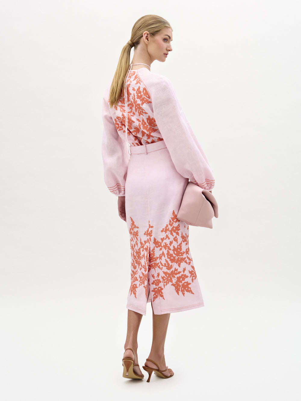 A woman faces away in a light pink floral blouse and the SVT DESIGNS Layan Skirt, paired with tan high-heeled sandals. She holds a pink clutch, and her blond hair is tied back in a low ponytail.