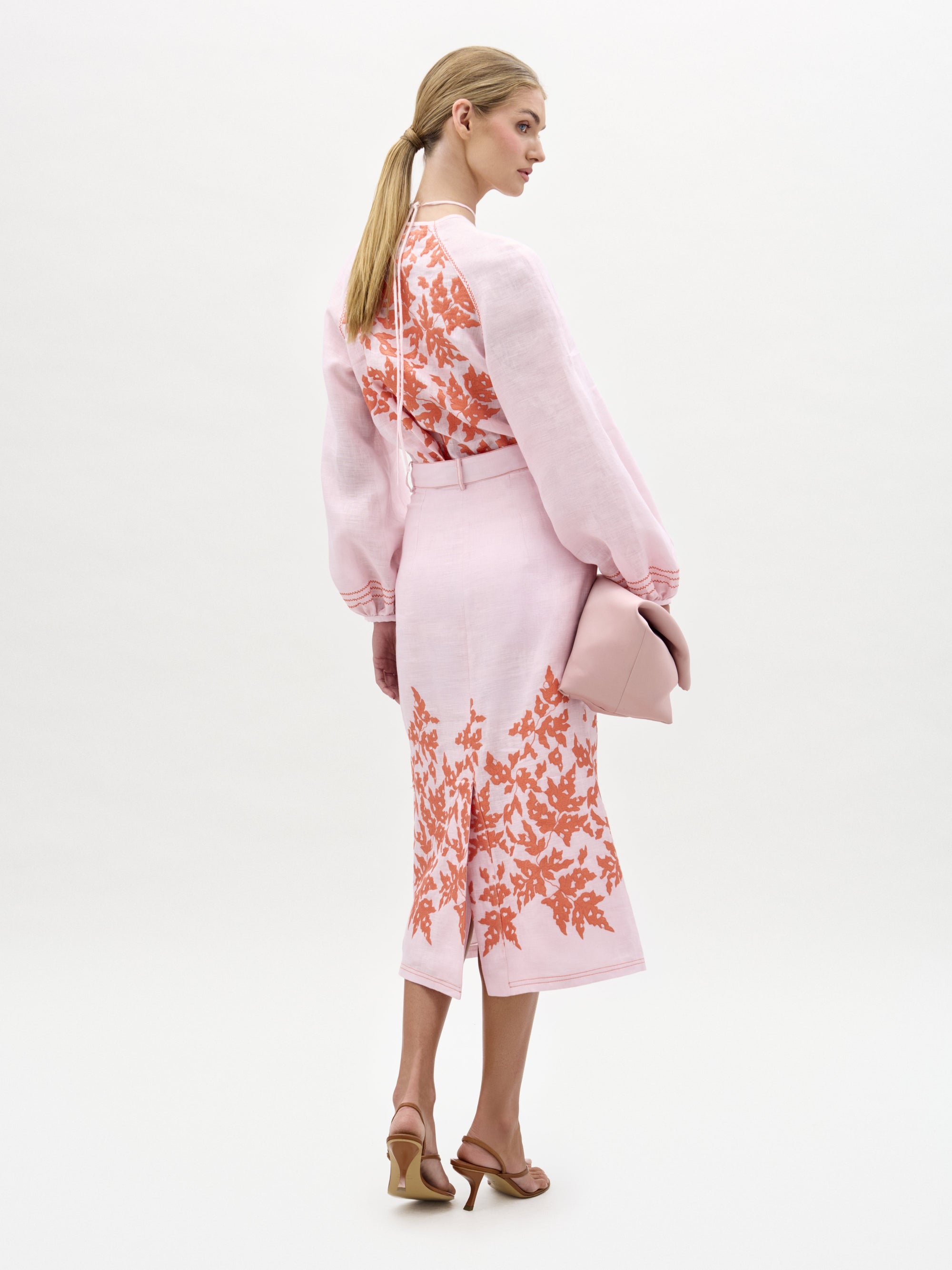 A woman faces away in a light pink floral blouse and the SVT DESIGNS Layan Skirt, paired with tan high-heeled sandals. She holds a pink clutch, and her blond hair is tied back in a low ponytail.