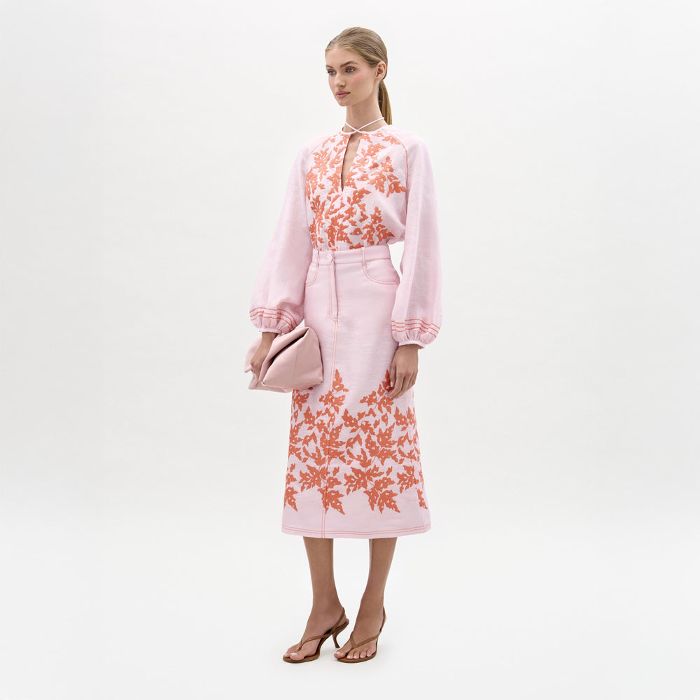 A woman stands against a plain white background wearing the SVT DESIGNS Layan Skirt, a structured midi skirt with orange floral embroidery made in Portugal, paired with a light pink blouse, pale pink clutch, and brown heeled sandals.