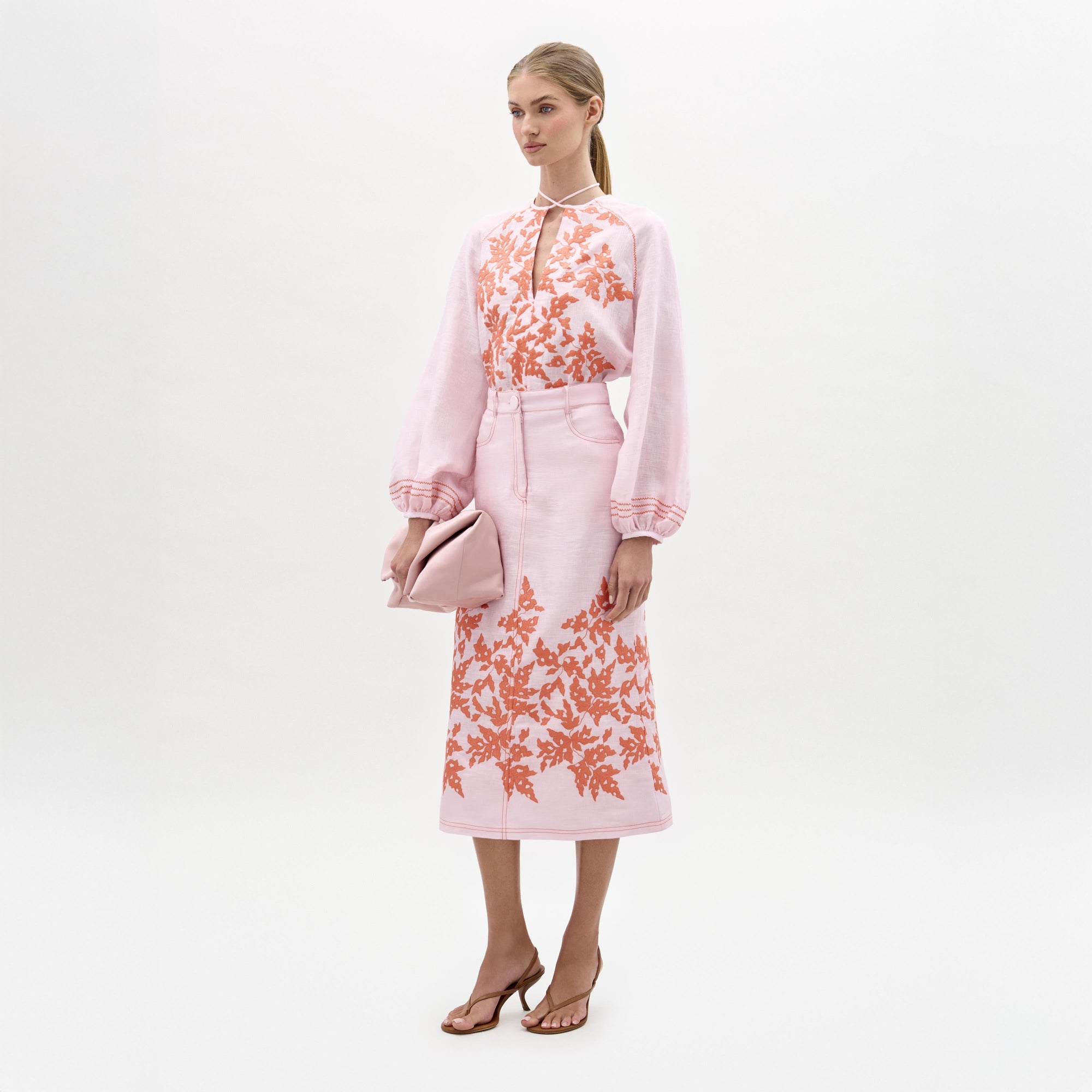 A woman stands against a plain white background wearing the SVT DESIGNS Layan Skirt, a structured midi skirt with orange floral embroidery made in Portugal, paired with a light pink blouse, pale pink clutch, and brown heeled sandals.