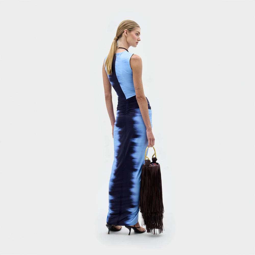 A woman faces away in the SVT DESIGNS Leni Dress, a form-fitting blue and black Italian-fabric midi with a tie-dye effect. She holds a large black fringed handbag, wears black heels, and has her hair in a ponytail.