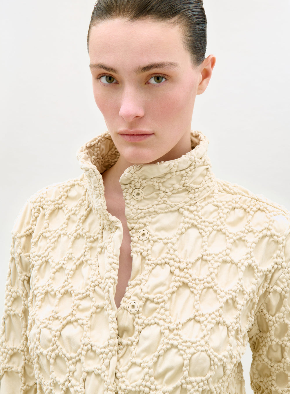 A person with fair skin and dark hair pulled back models the cream-colored, diamond-patterned Zalta Jacket with a high collar, looking directly at the camera against a plain light background.