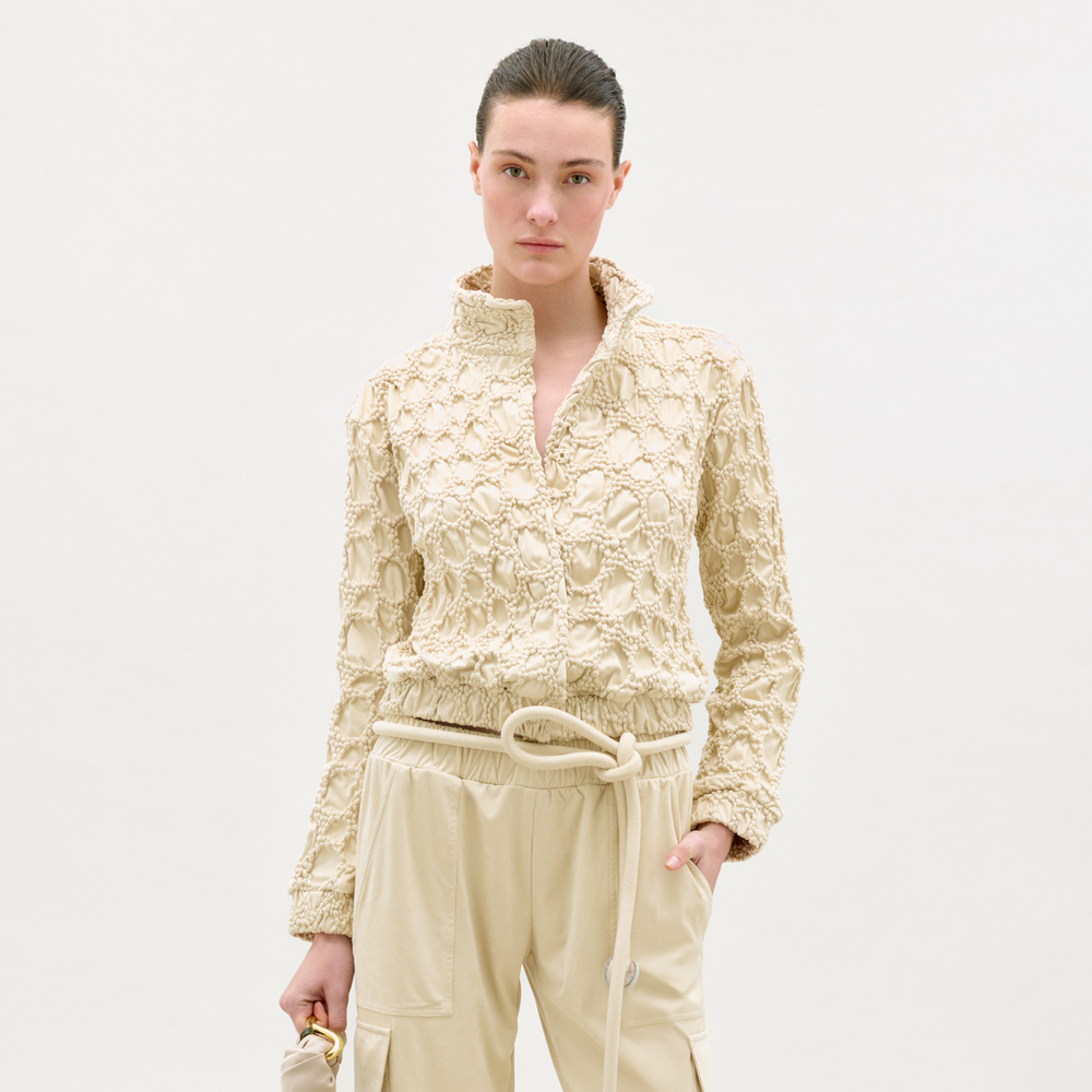 Wearing the Zalta Jacket, a textured cream-colored piece with a high collar, and light drawstring pants, a person stands against a plain background holding a small beige bag.