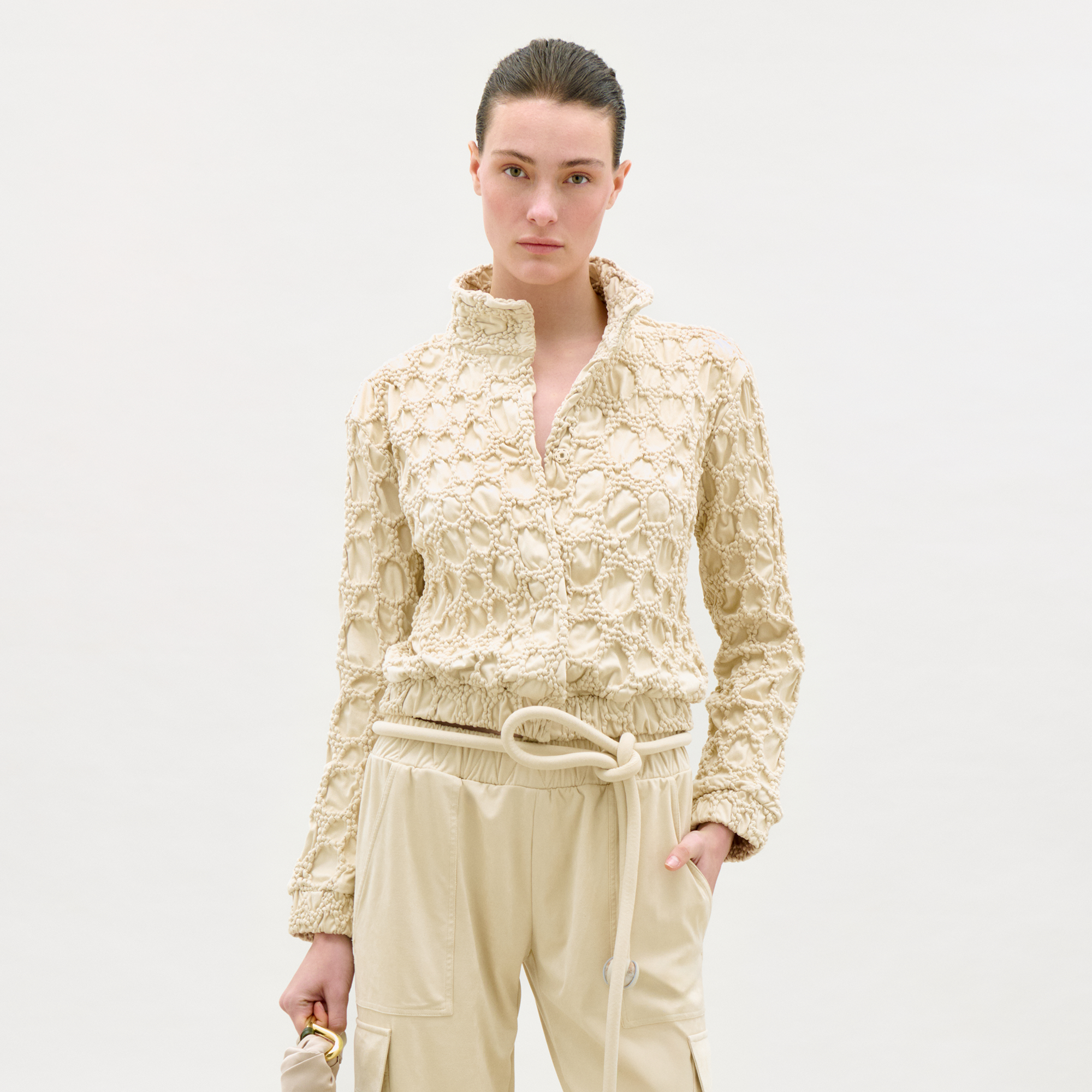 Wearing the Zalta Jacket, a textured cream-colored piece with a high collar, and light drawstring pants, a person stands against a plain background holding a small beige bag.