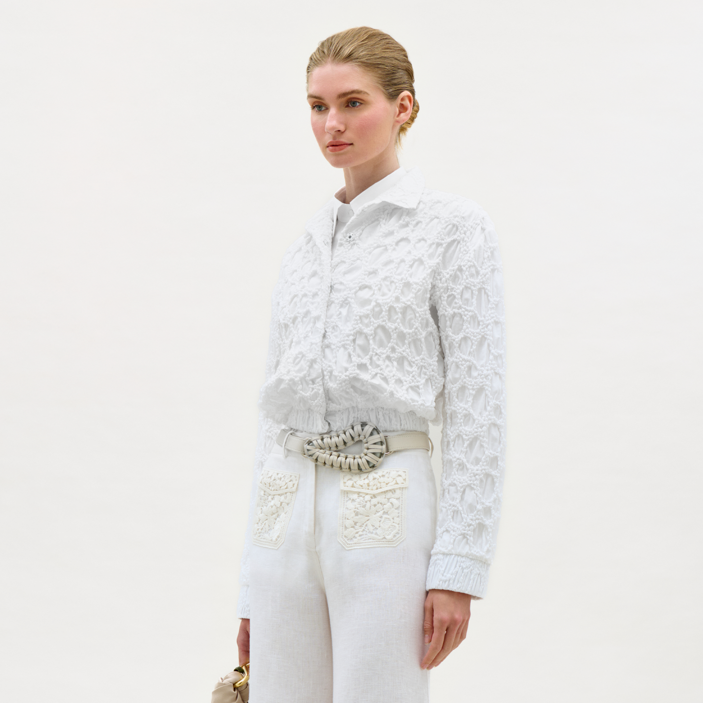 Woman wearing a white lace top and white pants with a belt on a light gray background