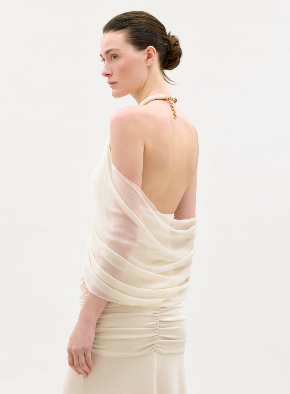 A woman with fair skin and dark hair in a low bun models the sheer, draped, off-shoulder Clio Top with a ruched skirt, standing sideways and gazing left against a plain light background.