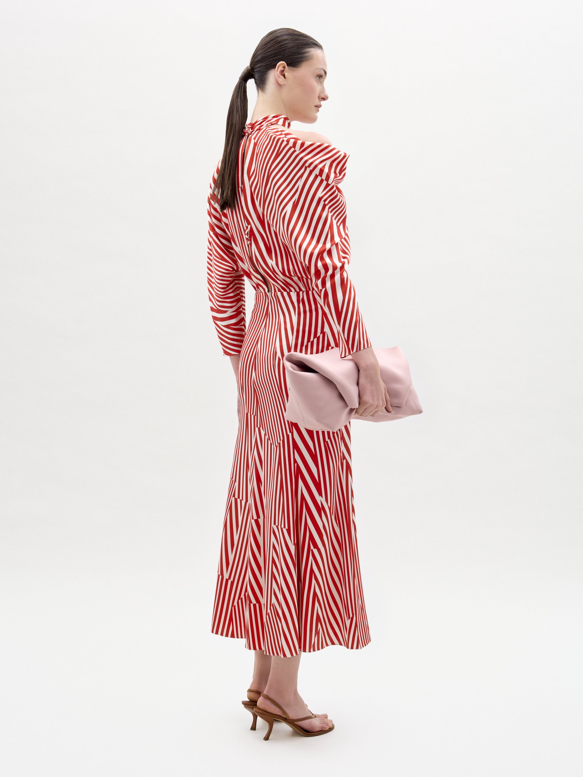 A woman stands in profile wearing the Lisa Dress by SVT DESIGNS, a long-sleeve midi dress with red and white stripes. She holds a large light pink clutch, wears brown heeled sandals, and has her hair in a low ponytail against a white background.
