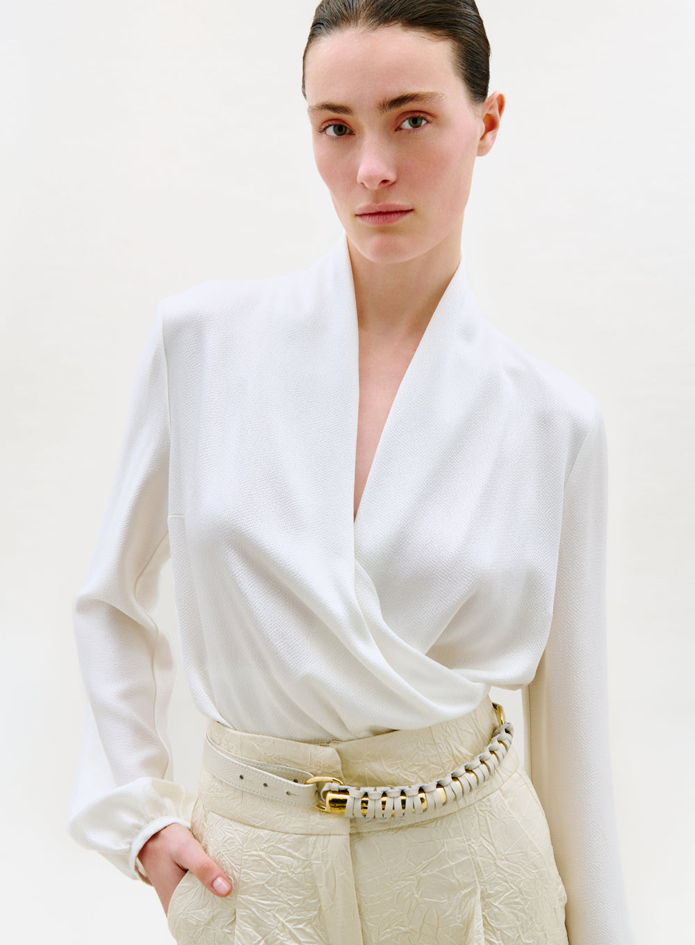 A woman with dark hair pulled back models the Tea Blouse—a white wrap top—paired with high-waisted, textured cream pants and a wide belt with gold accents, standing against a plain light background.