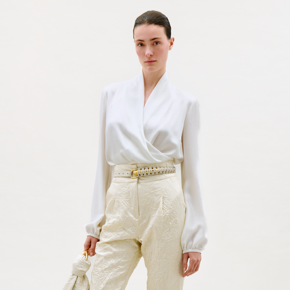 Woman wearing a white blouse and cream pants against a plain background