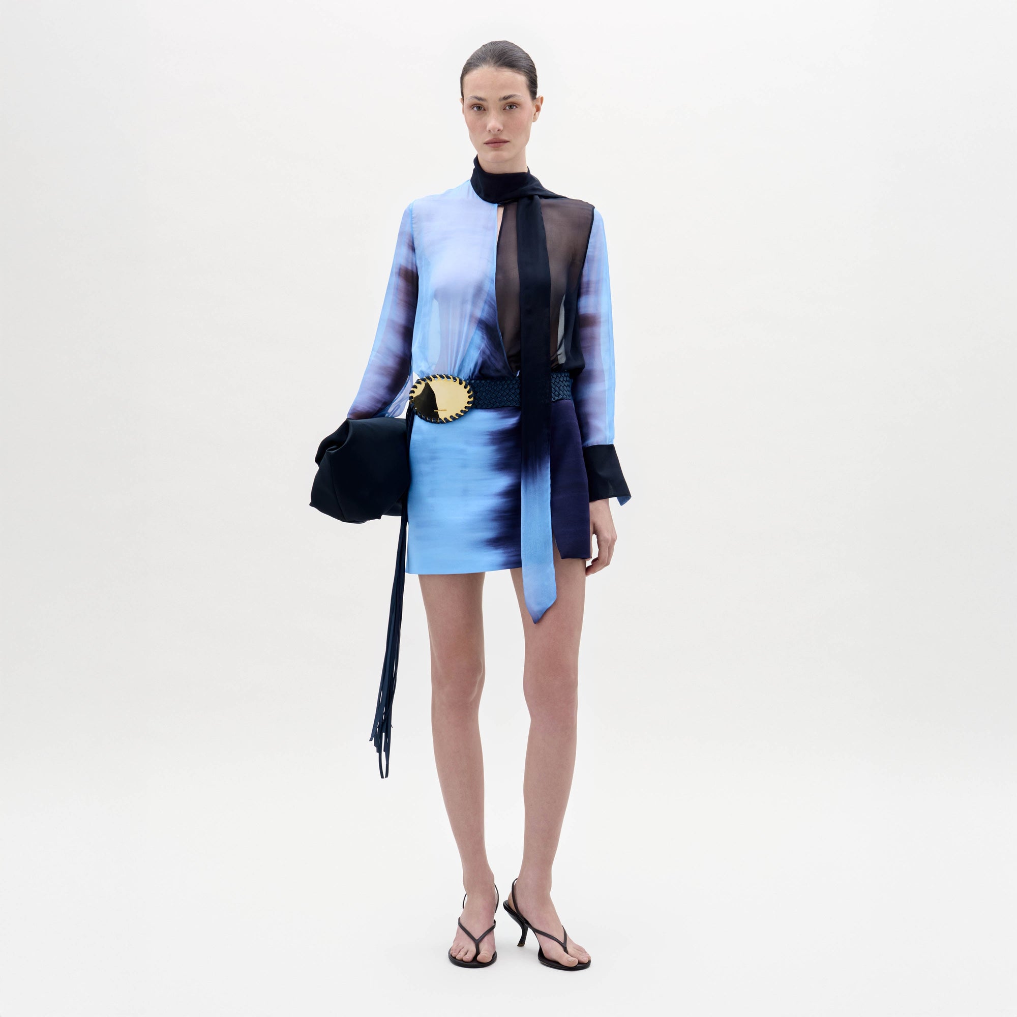 A woman stands against a white background wearing the Imelda Dress, styled with black sandals, a black bag, and a belt featuring a large gold decorative buckle.