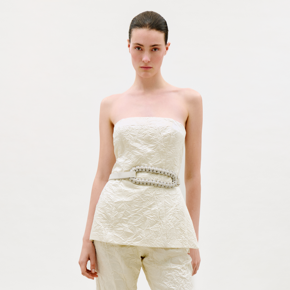 A woman with dark hair in a low ponytail wears the Astor Top, a strapless, textured off-white piece paired with matching pants and a wide beaded belt. She stands against a plain light background, looking directly at the camera.