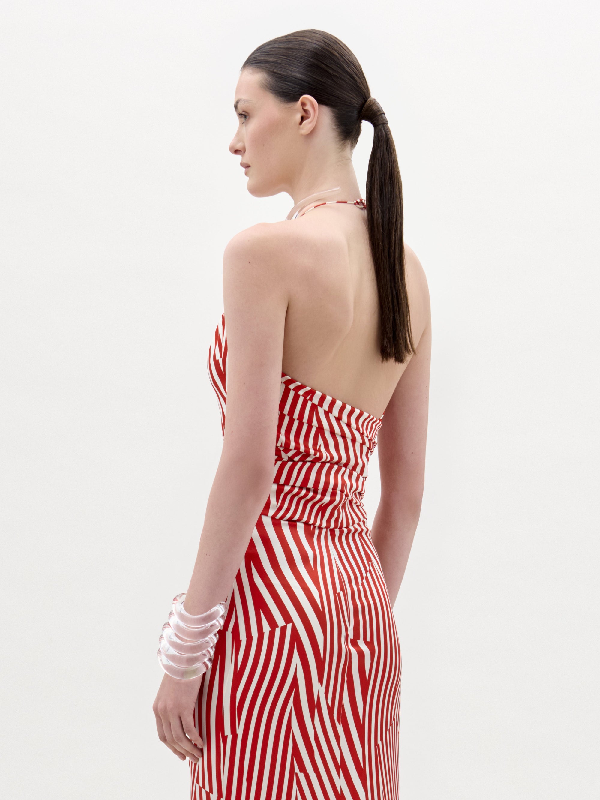 A woman with straight dark hair in a low ponytail wears the Carmela Blouse by SVT DESIGNS, featuring red and white stripes and a chic halter neckline. She stands in profile against a plain white background, wearing clear bangle bracelets.