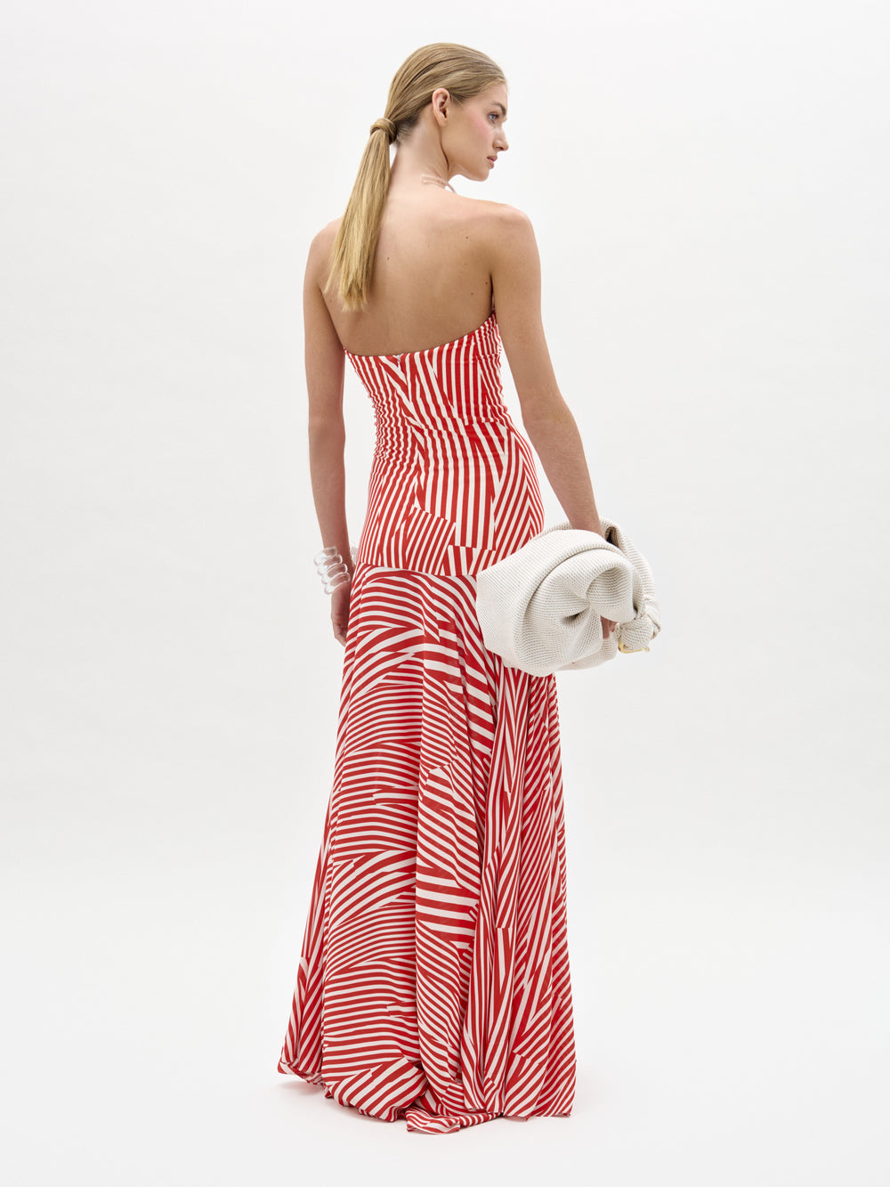 A woman with long blond hair models the Muriel Dress by SVT DESIGNS—a red and white strapless maxi dress with diagonal stripes and a fitted bodice—standing away from the camera, holding an off-white clutch against a white background.