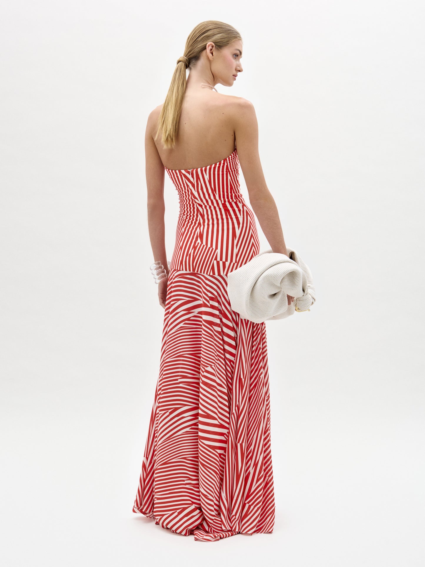 A woman with long blond hair models the Muriel Dress by SVT DESIGNS—a red and white strapless maxi dress with diagonal stripes and a fitted bodice—standing away from the camera, holding an off-white clutch against a white background.