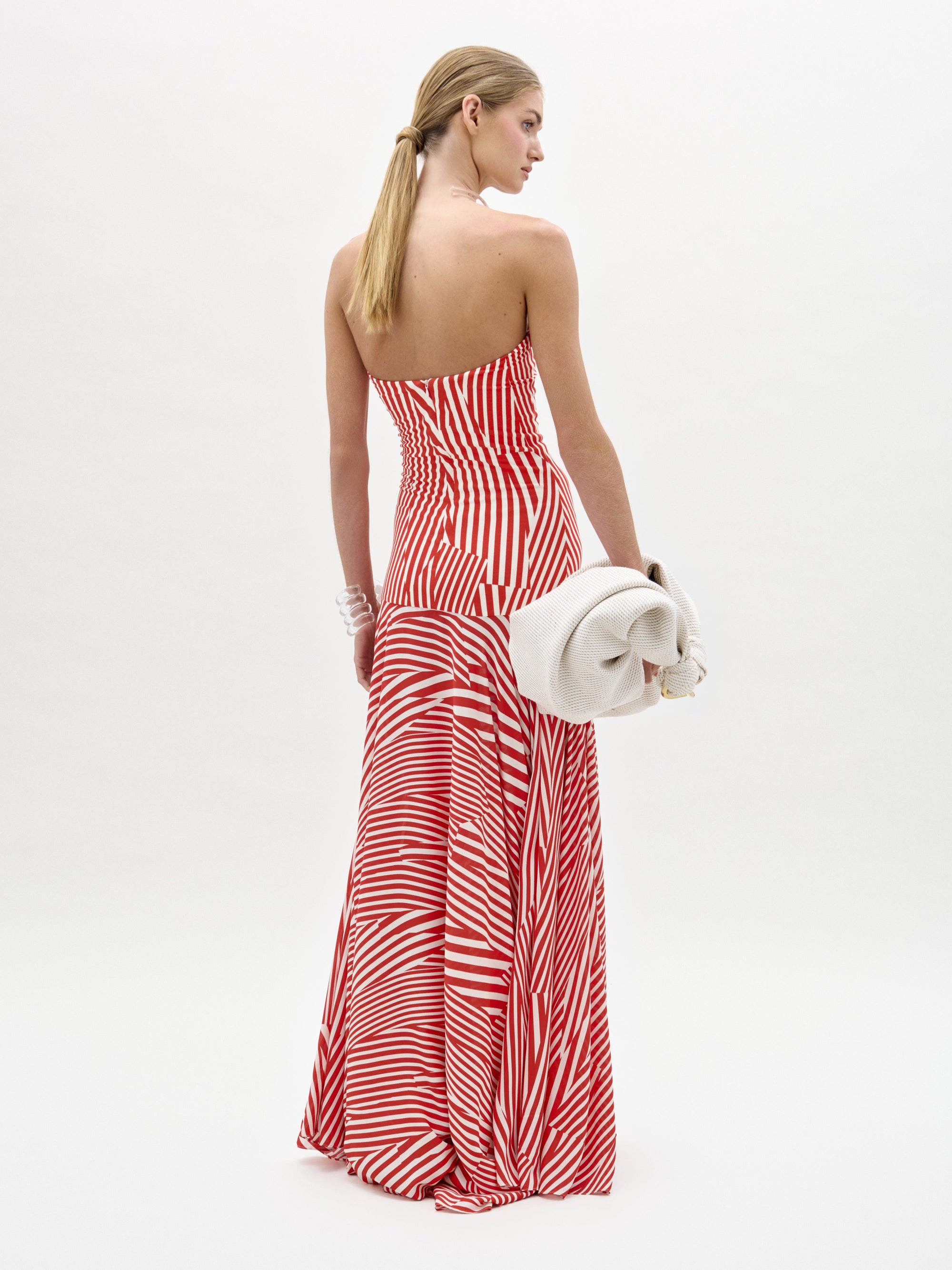 A woman with long blond hair models the Muriel Dress by SVT DESIGNS—a red and white strapless maxi dress with diagonal stripes and a fitted bodice—standing away from the camera, holding an off-white clutch against a white background.