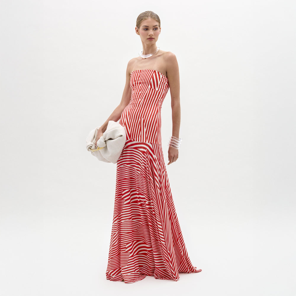 A woman poses against a white background in the SVT DESIGNS Muriel Dress, a strapless red and white striped maxi with a fitted bodice. She accessorizes with a pearl necklace, bracelets, and holds a white clutch bag.