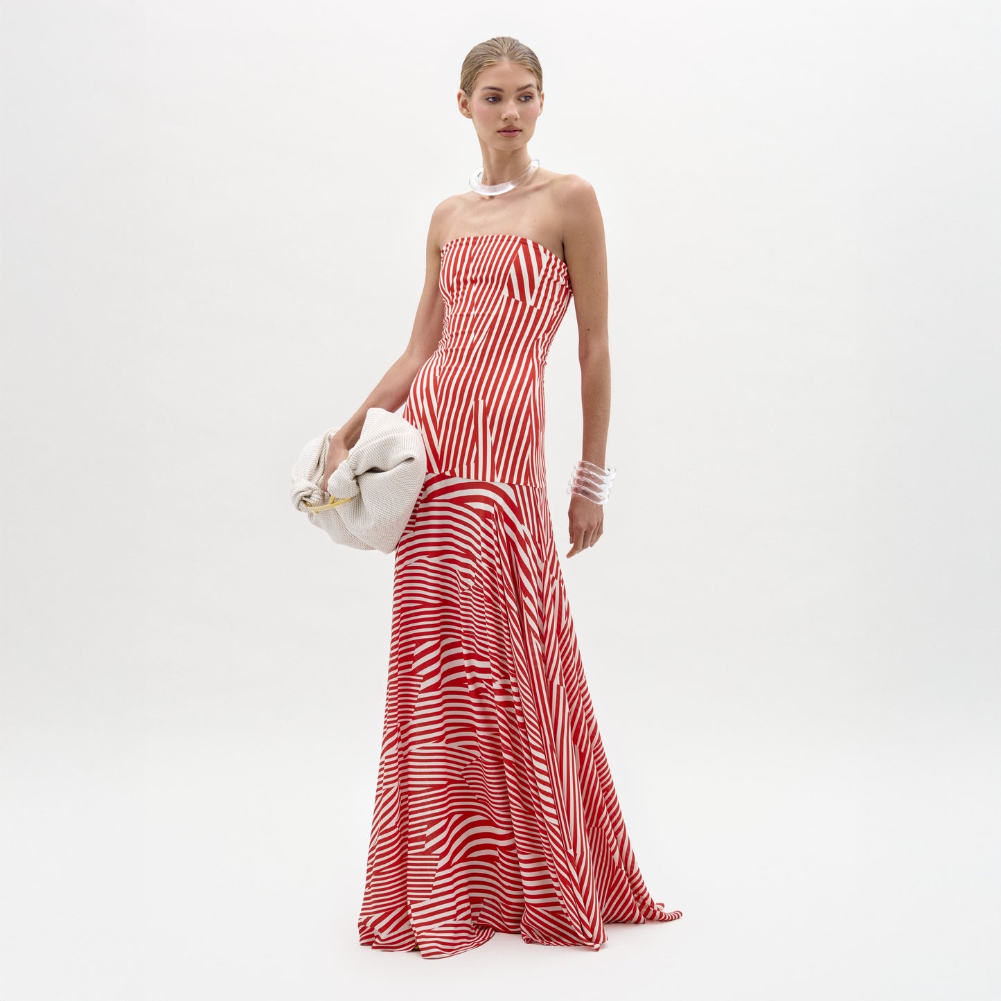 A woman poses against a white background in the SVT DESIGNS Muriel Dress, a strapless red and white striped maxi with a fitted bodice. She accessorizes with a pearl necklace, bracelets, and holds a white clutch bag.