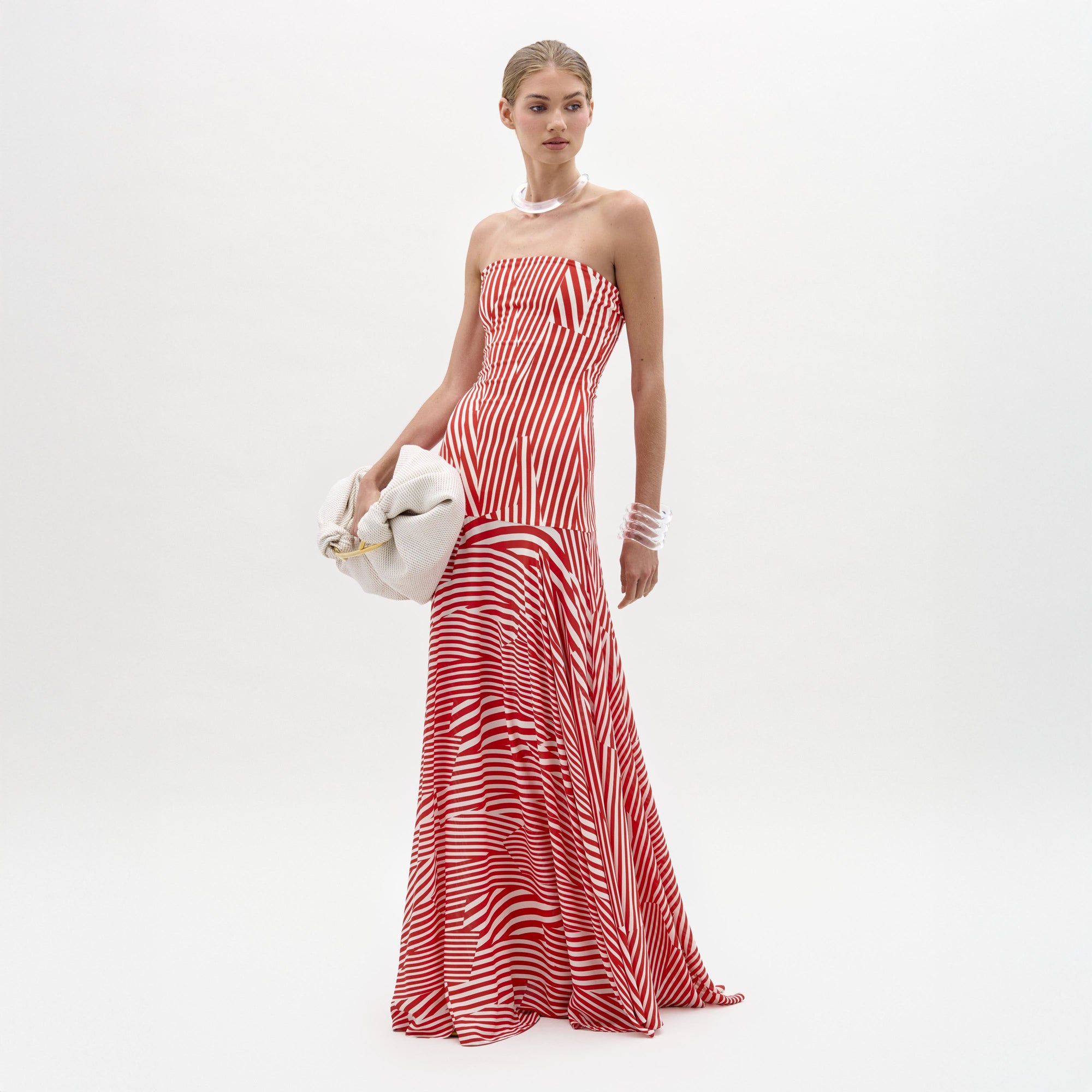 A woman poses against a white background in the SVT DESIGNS Muriel Dress, a strapless red and white striped maxi with a fitted bodice. She accessorizes with a pearl necklace, bracelets, and holds a white clutch bag.
