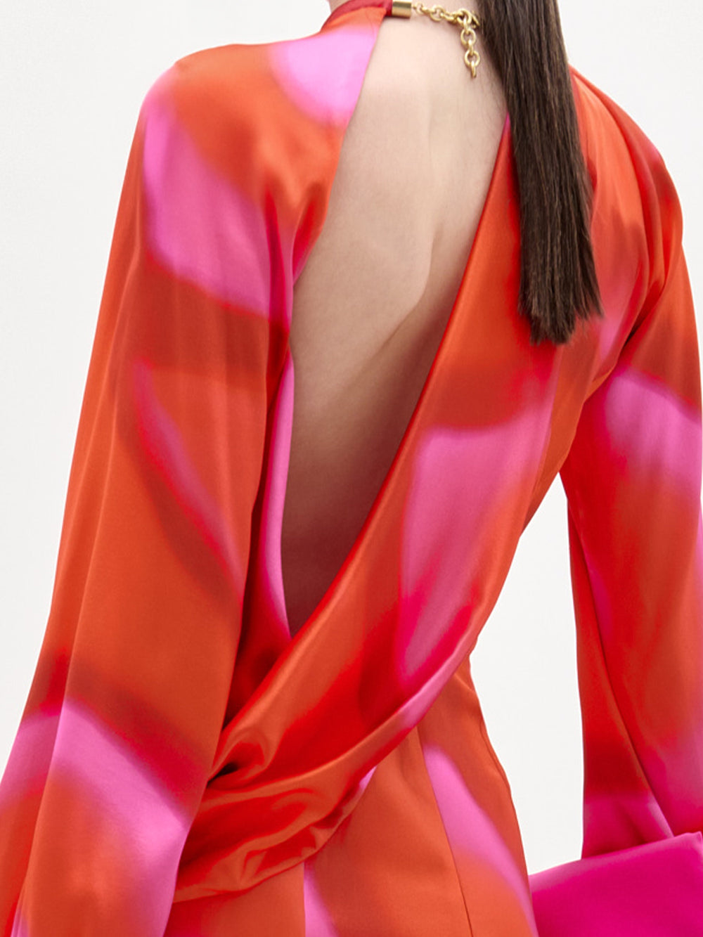 A person with long dark hair wears the SVT DESIGNS Muna Dress, a red and pink patterned silk midi with an open back secured by a gold clasp at the neck. The image highlights the dress's upper back and shoulder details.