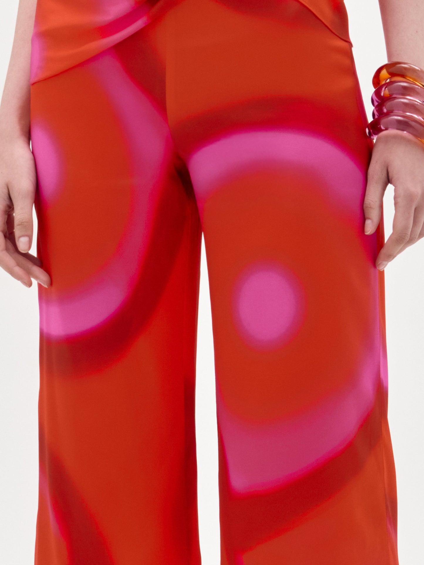 A person stands with arms relaxed at their sides, wearing SVT DESIGNS’ Andie Pant, a bright orange and pink high-waisted wide-leg style with a bold circular pattern crafted from fine Italian fabric, plus two translucent stacked bracelets on their right wrist.