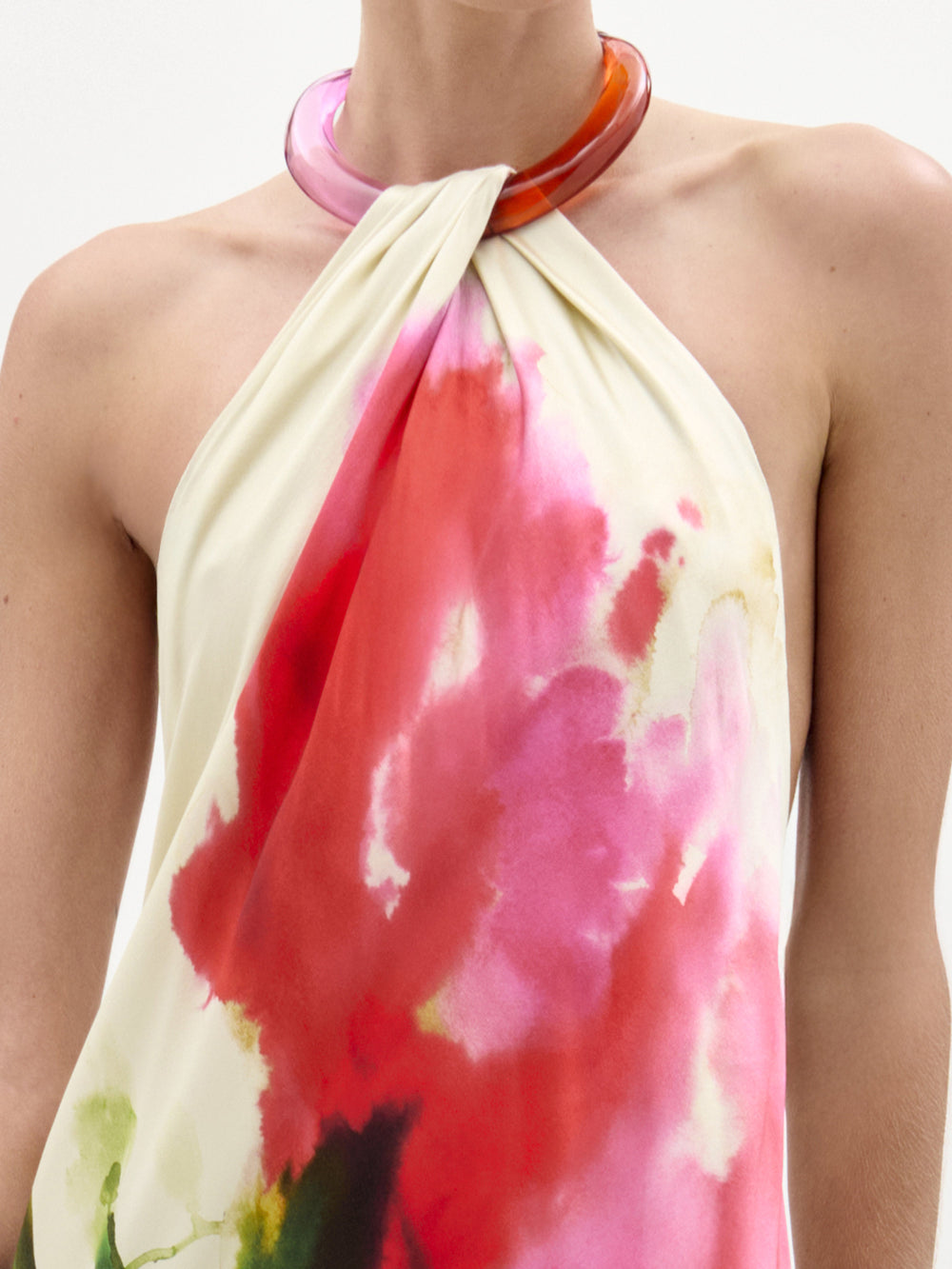 A person wears the SVT DESIGNS Jesse Dress, a sleeveless halter-neck style with a pink and red watercolor pattern, paired with a thick translucent reddish-pink necklace. The image is cropped to show shoulders and upper chest.