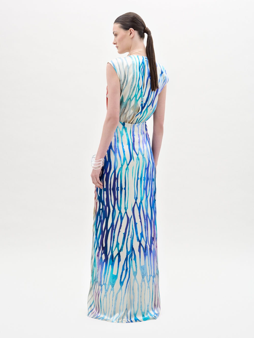 A woman with a low ponytail models the Samaira Dress by SVT DESIGNS, a sleeveless maxi with a ruched waist and blue, purple, and white abstract pattern. She wears a white bracelet on her left wrist against a plain white background.